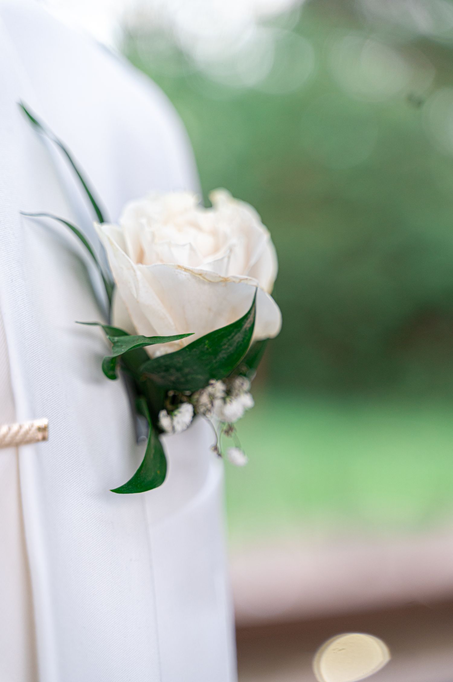 White rose boutonniere with green leaves pinned to grooms white wedding suit lapel.