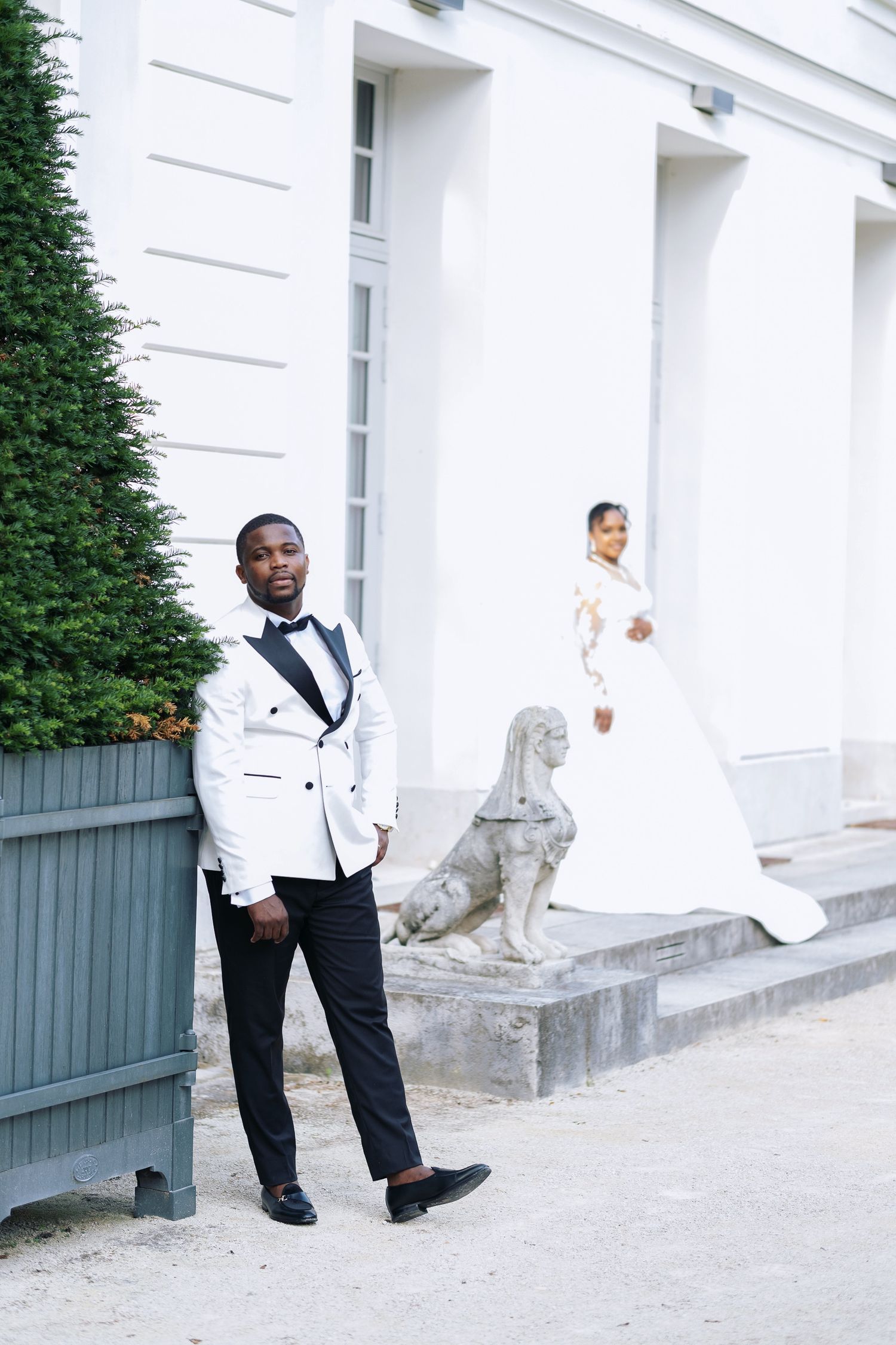 Elegant groom in white tuxedo poses outdoors while bride in lace wedding gown stands in background.