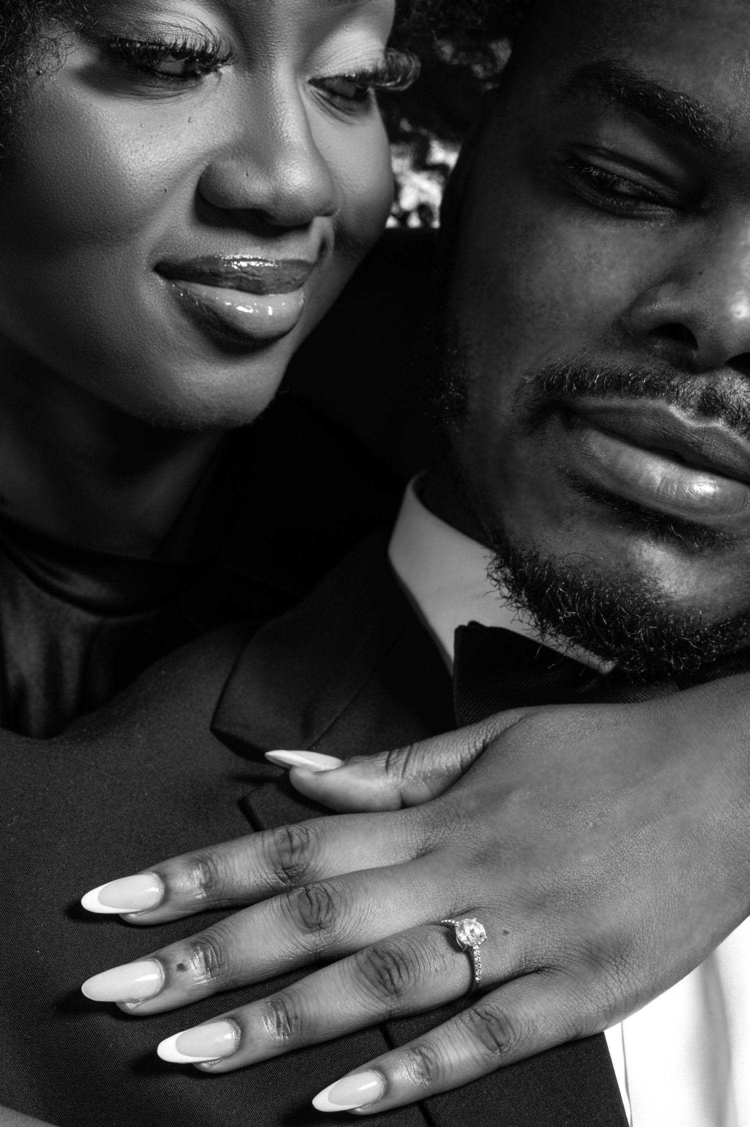 Black couple in elegant attire, woman showing diamond engagement ring with white manicured nails, black and white photo.