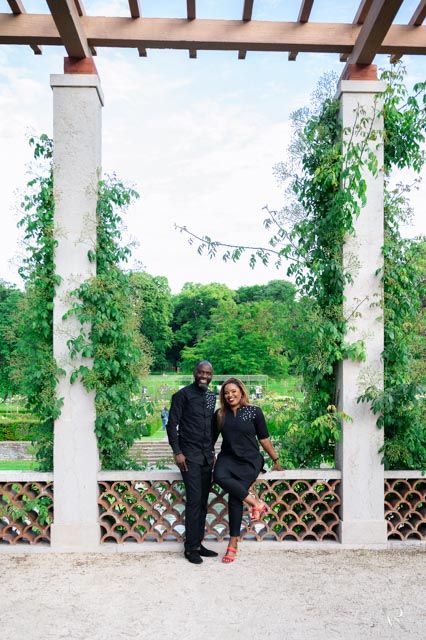 Couple in matching black outfits posing under a vine-covered pergola in a lush garden setting.