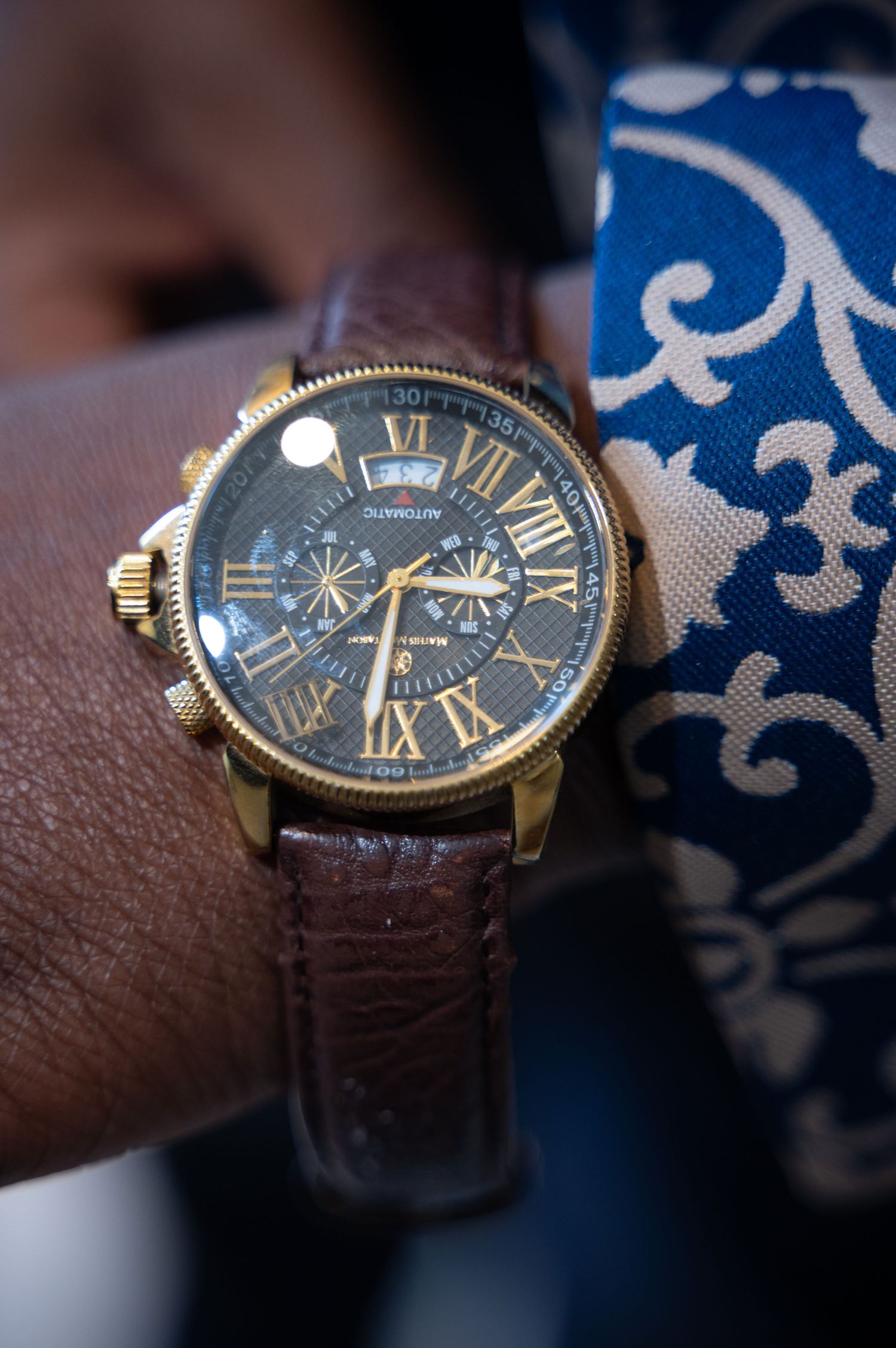 Close-up of a gold chronograph watch with brown leather strap on a groom's wrist at a wedding.