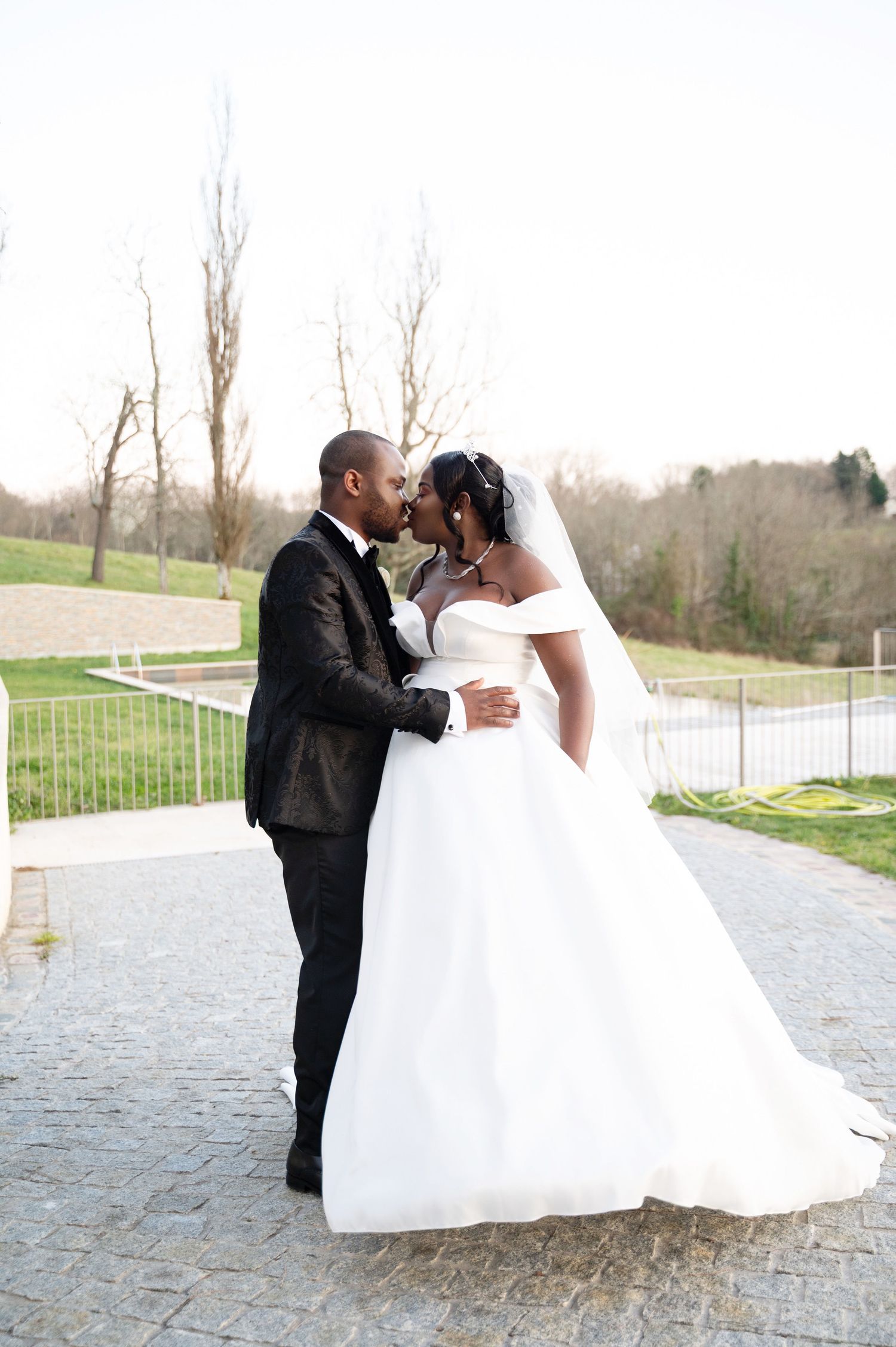 A Black bride and groom share a romantic kiss outdoors, with the bride in a full white ballgown and veil.