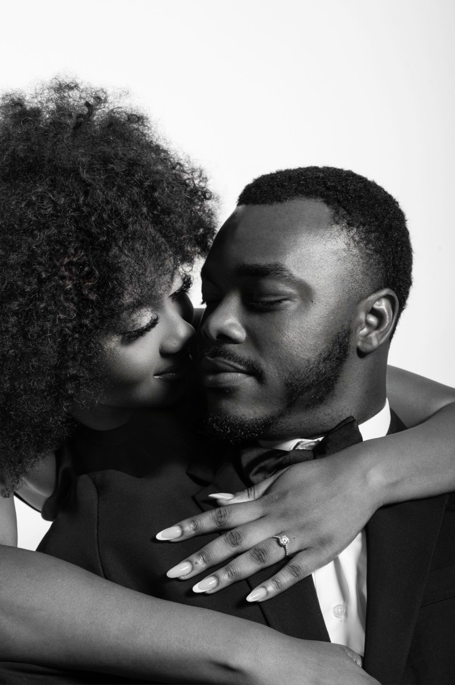 Black and white photo of a couple in formal wear sharing an intimate embrace, woman wearing an engagement ring.