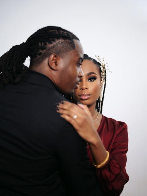 Elegant Black couple in formal attire share a romantic embrace, woman wearing burgundy dress and gold hair accessory.