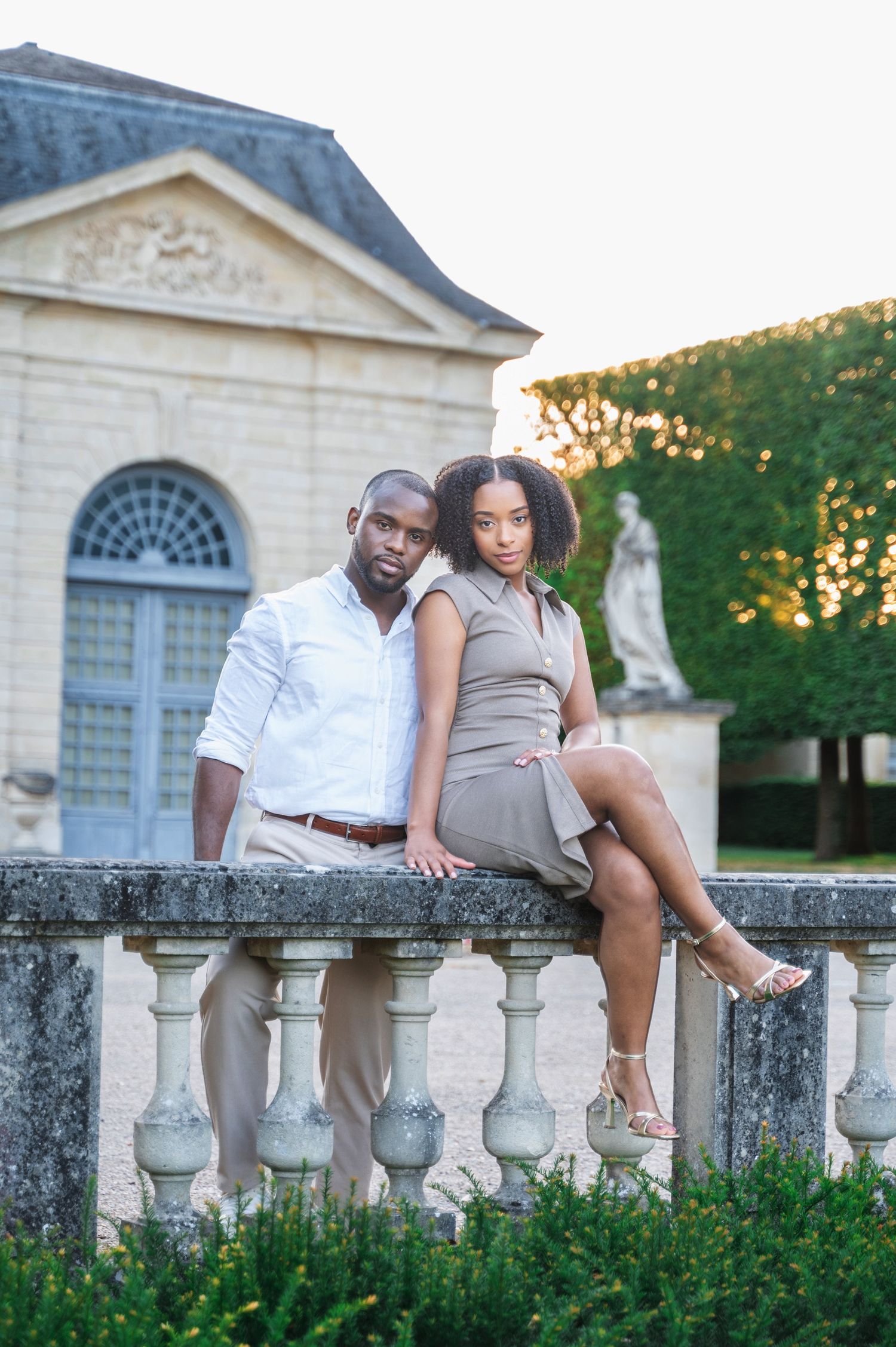 An elegant Black couple poses together on a stone balcony railing with a classic French chateau in the background.