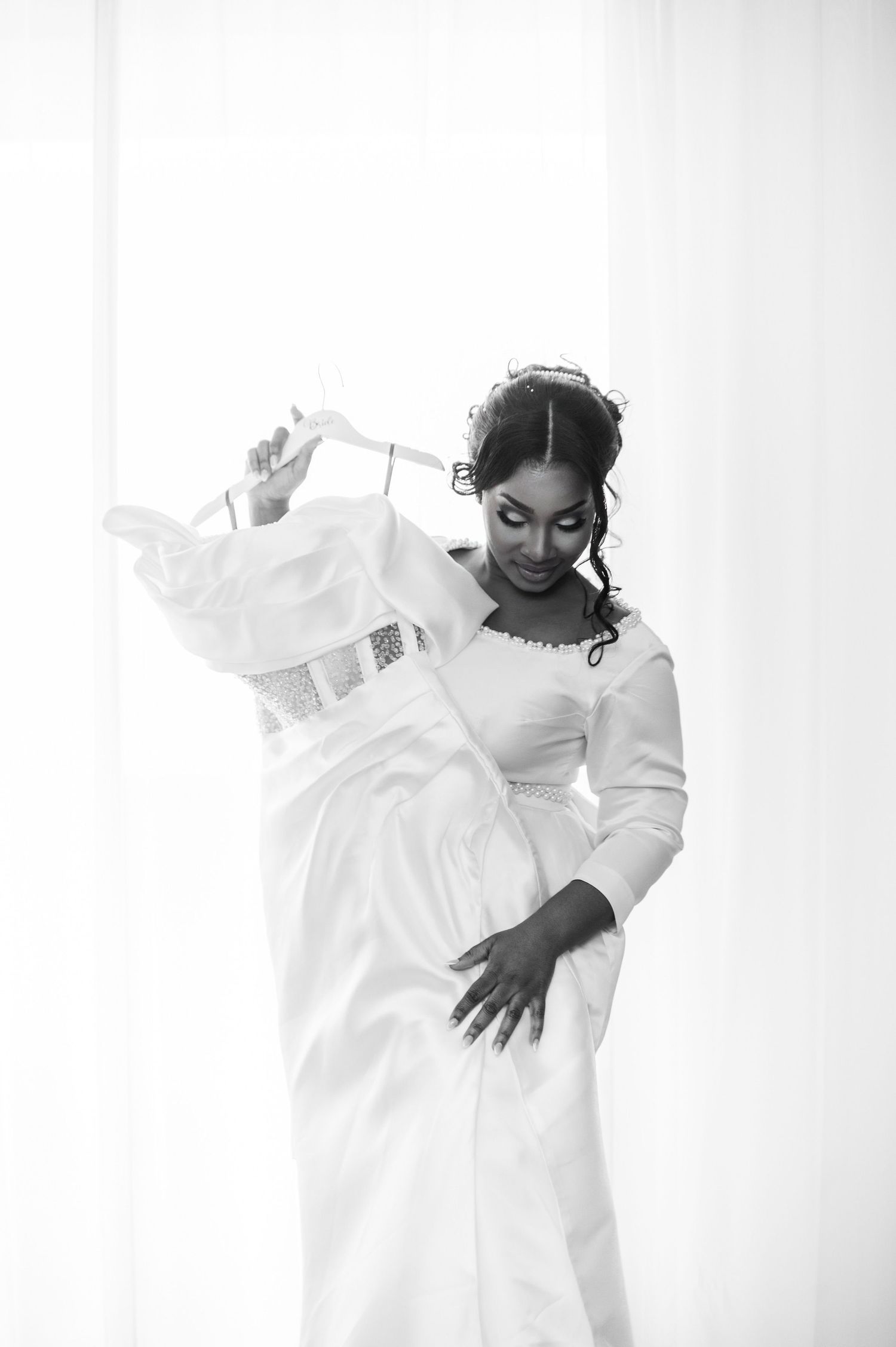 Black and white portrait of a bride holding her elegant wedding gown near a bright window.