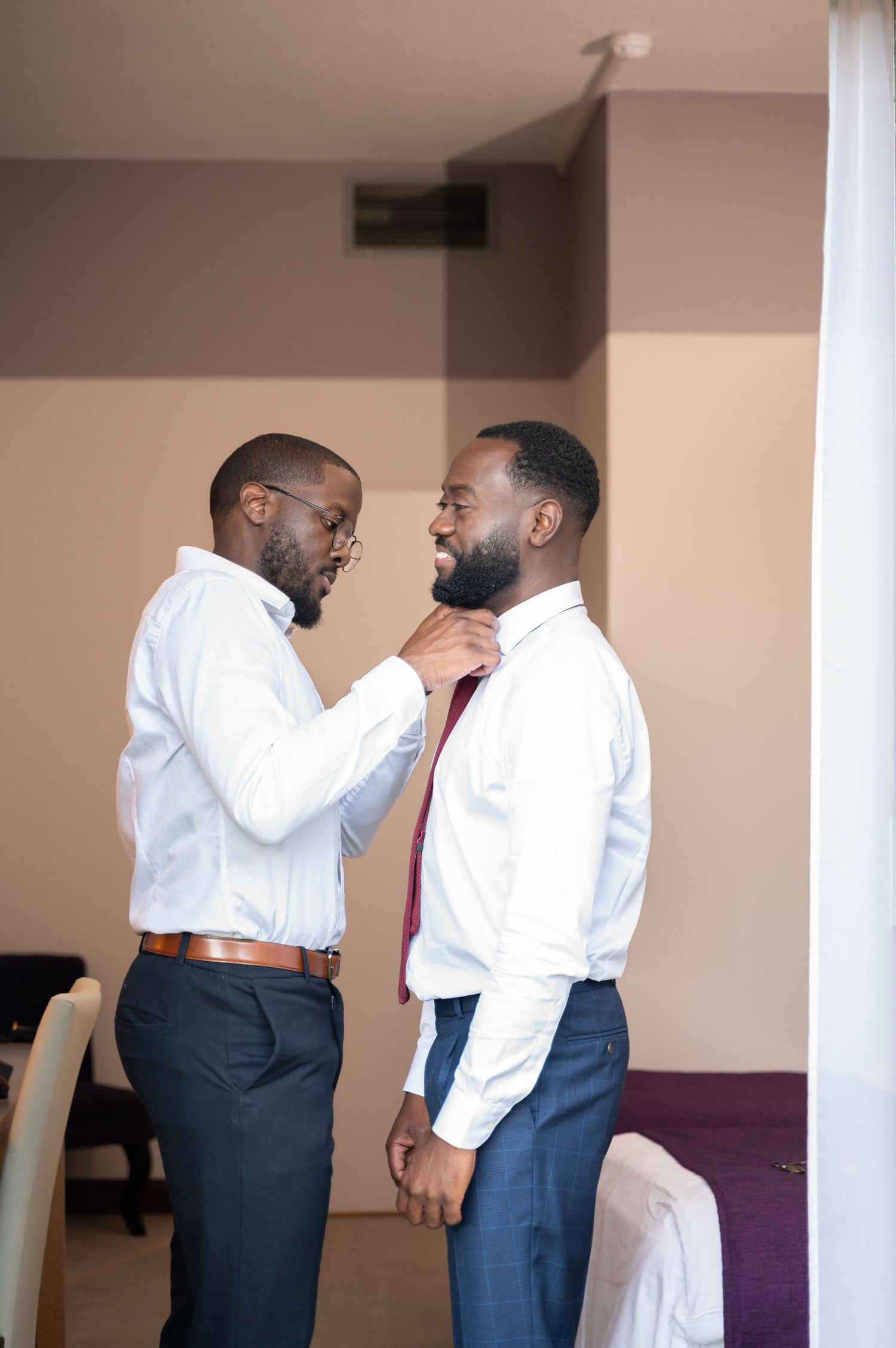 Groomsman helping groom adjust burgundy tie while both wear white dress shirts and navy blue pants.