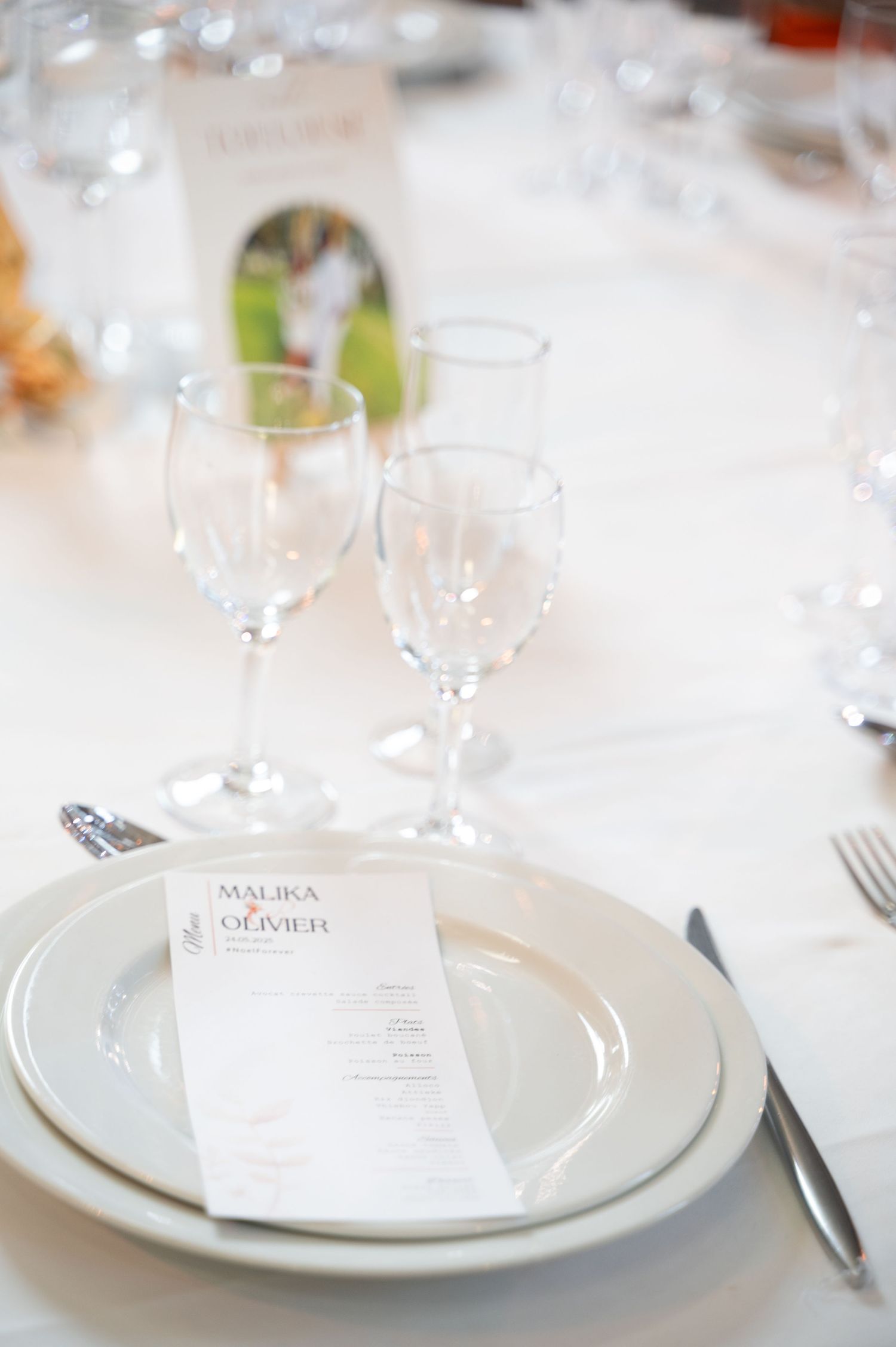 Wedding place setting with Malika and Olivier menu card on white plates with crystal wine glasses.