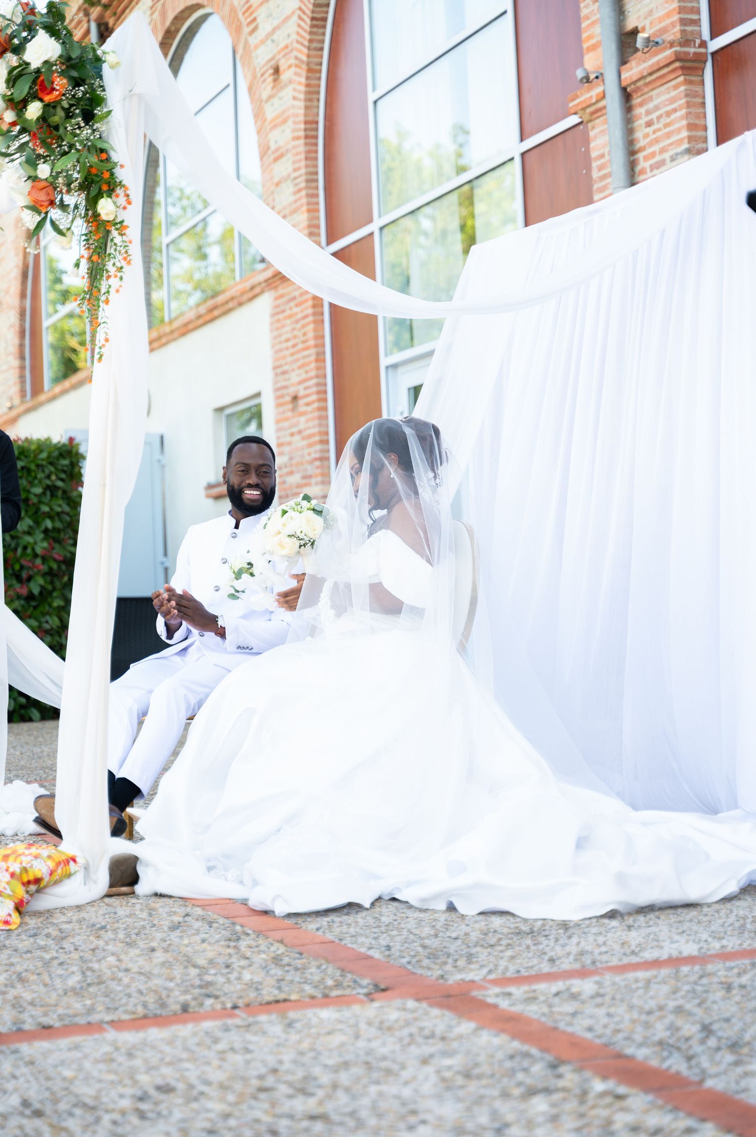 Laughing groom holds bride's hand under white draped canopy at outdoor wedding ceremony.