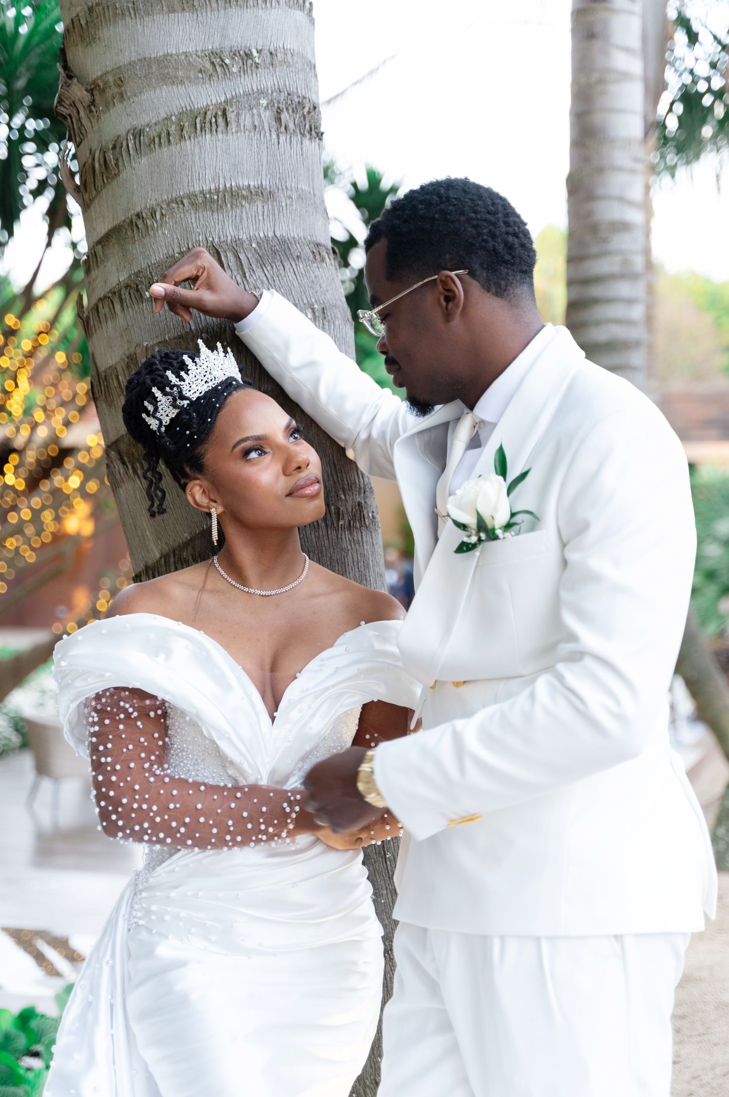 Bride in pearl-embellished gown and crown gazes at groom in white suit near palm tree.