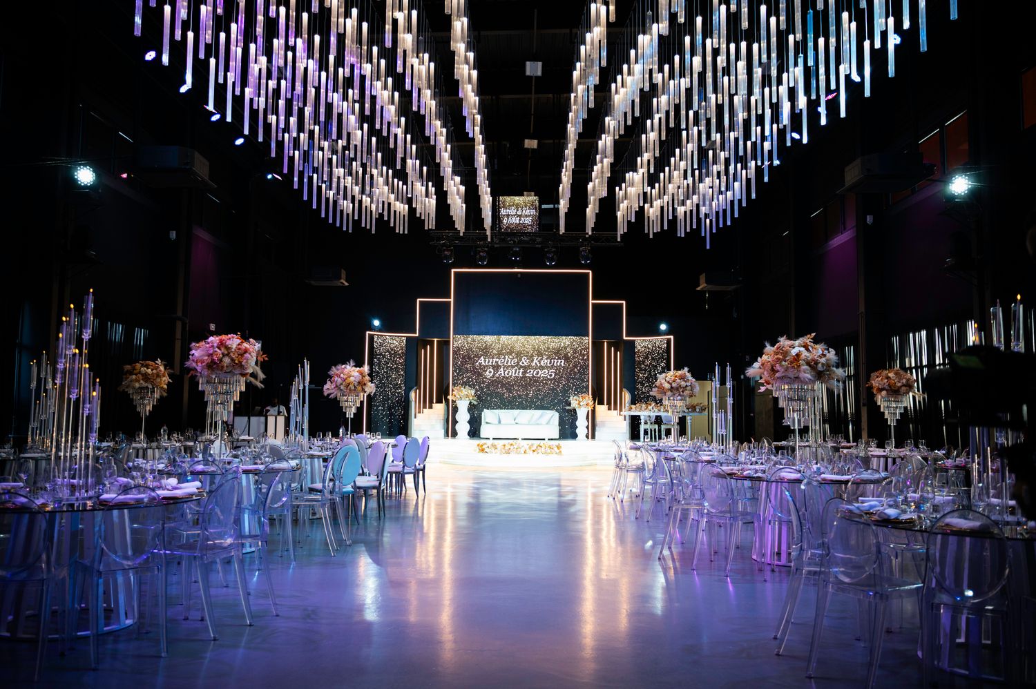 Elegant wedding reception hall with hanging light installations, ghost chairs, and pink floral centerpieces.