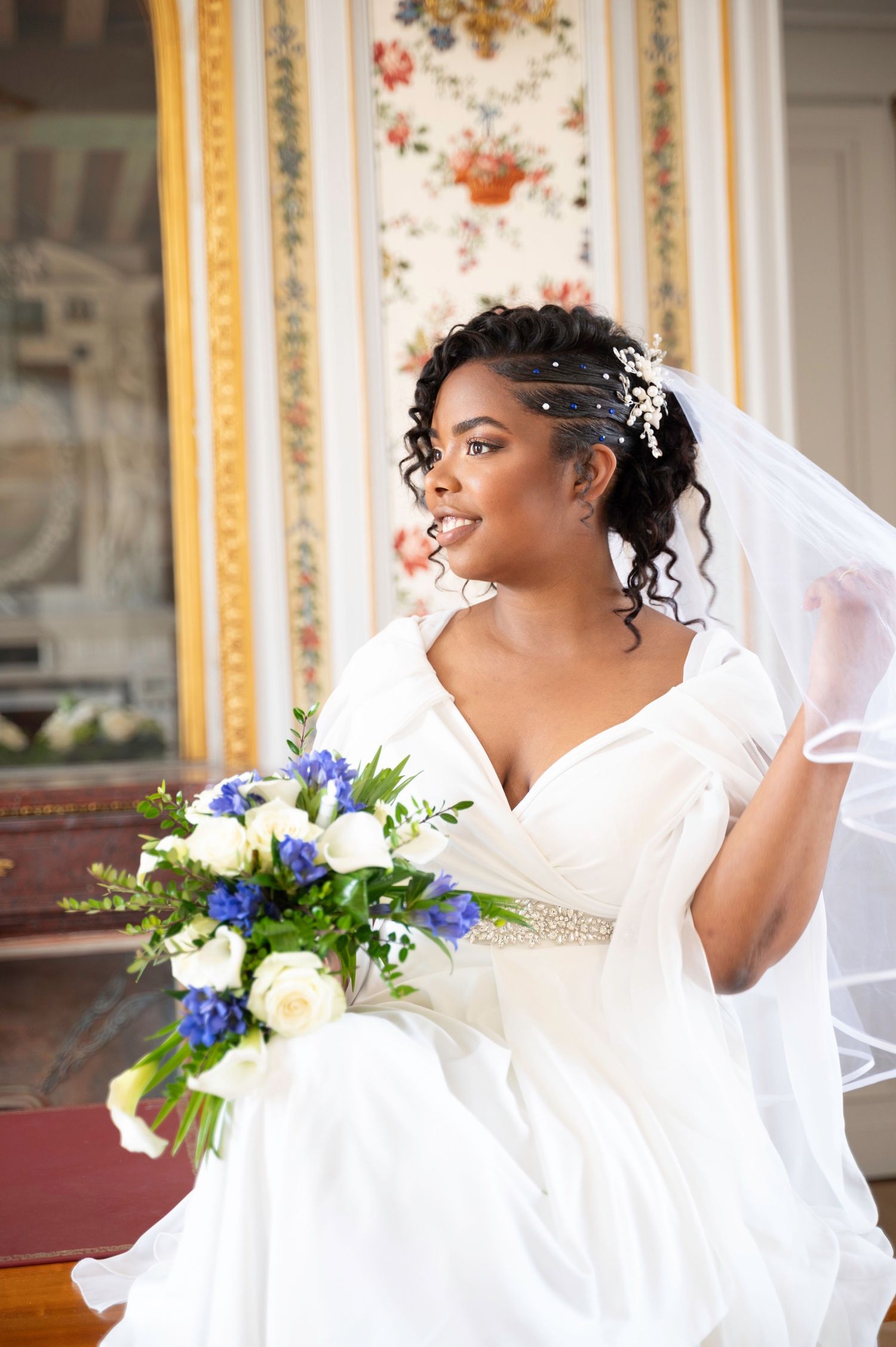 Beautiful Black bride in white gown holds blue and white bouquet inside elegant venue.