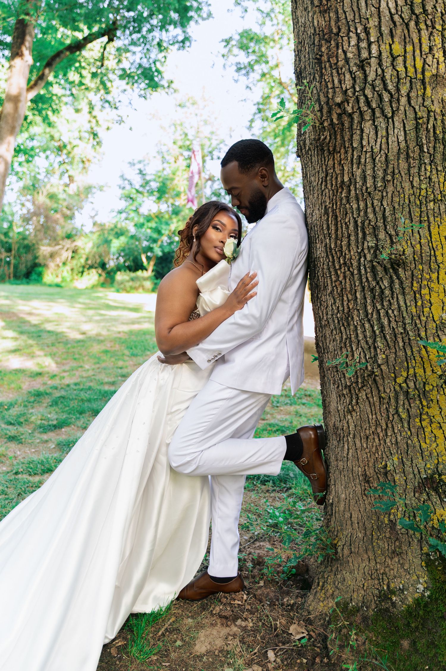 Romantic bride and groom in white wedding attire sharing an intimate embrace against a large tree.