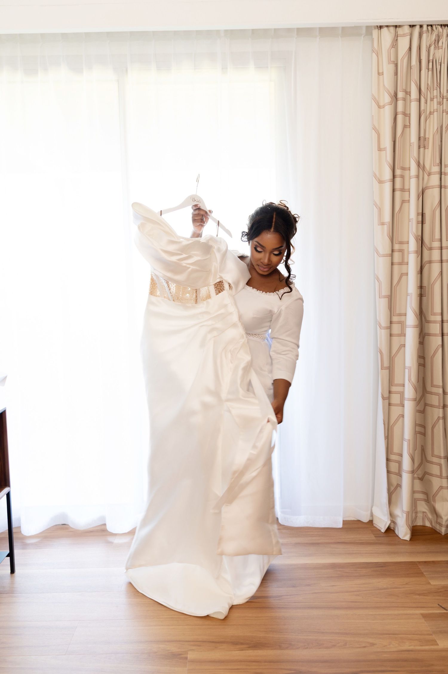 Bride holding elegant white wedding gown near bright window in hotel room.