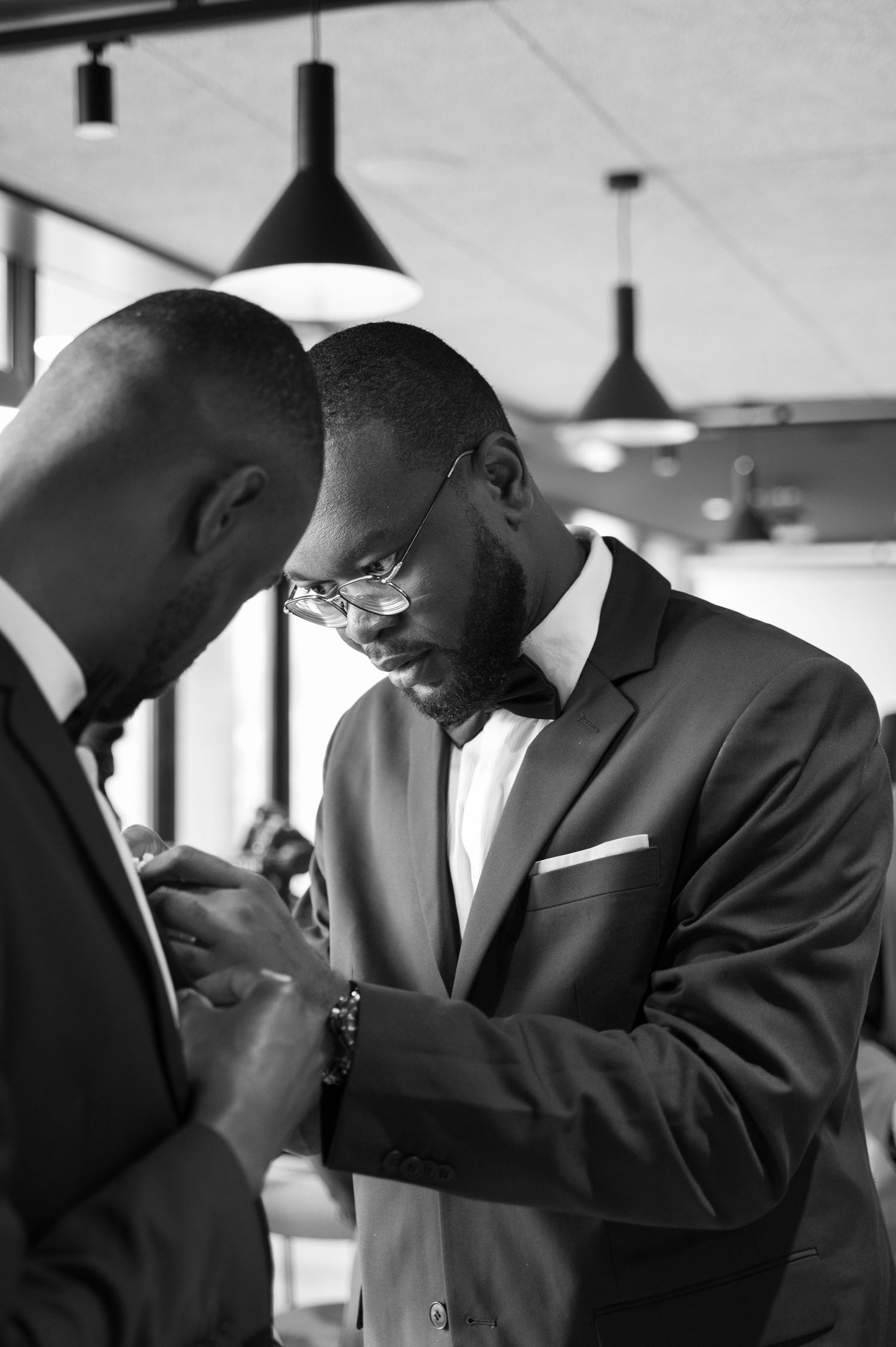 Black and white photo of two Black men in suits helping each other with wedding day preparations indoors.