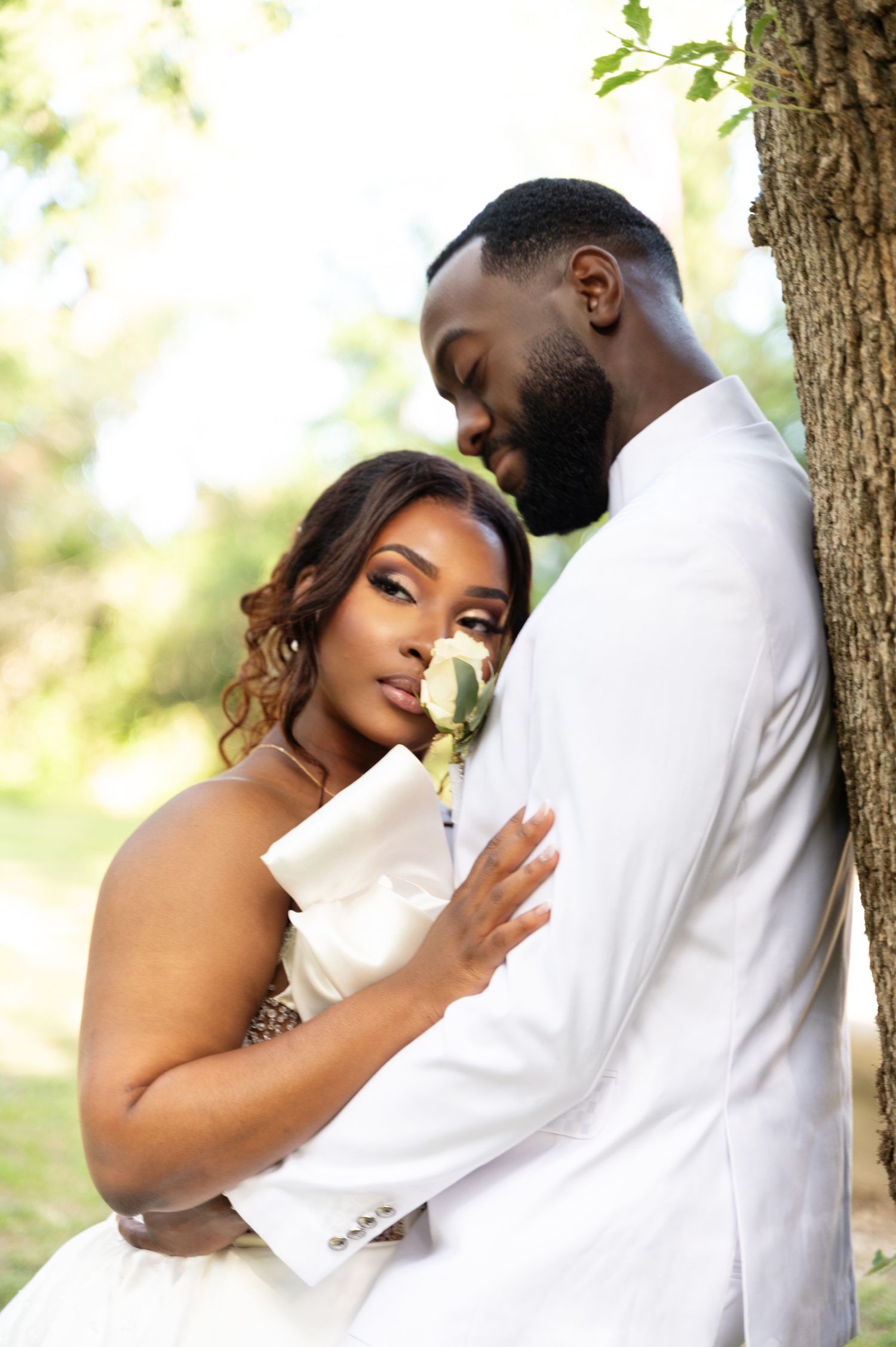 Black couple in white wedding attire embrace romantically against a tree in a sunlit outdoor setting.