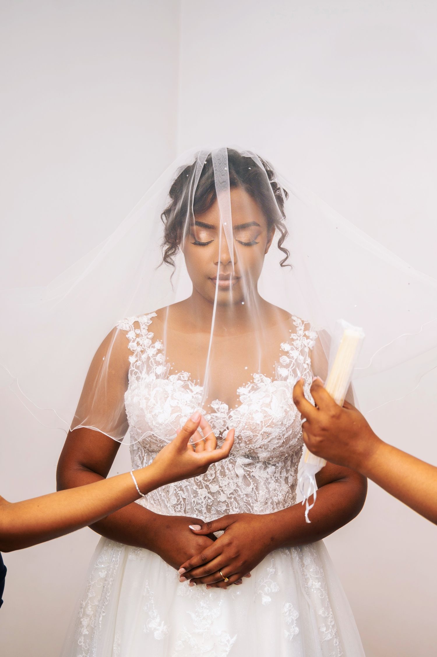 Bride standing with eyes closed under a veil while attendants adjust her wedding dress.