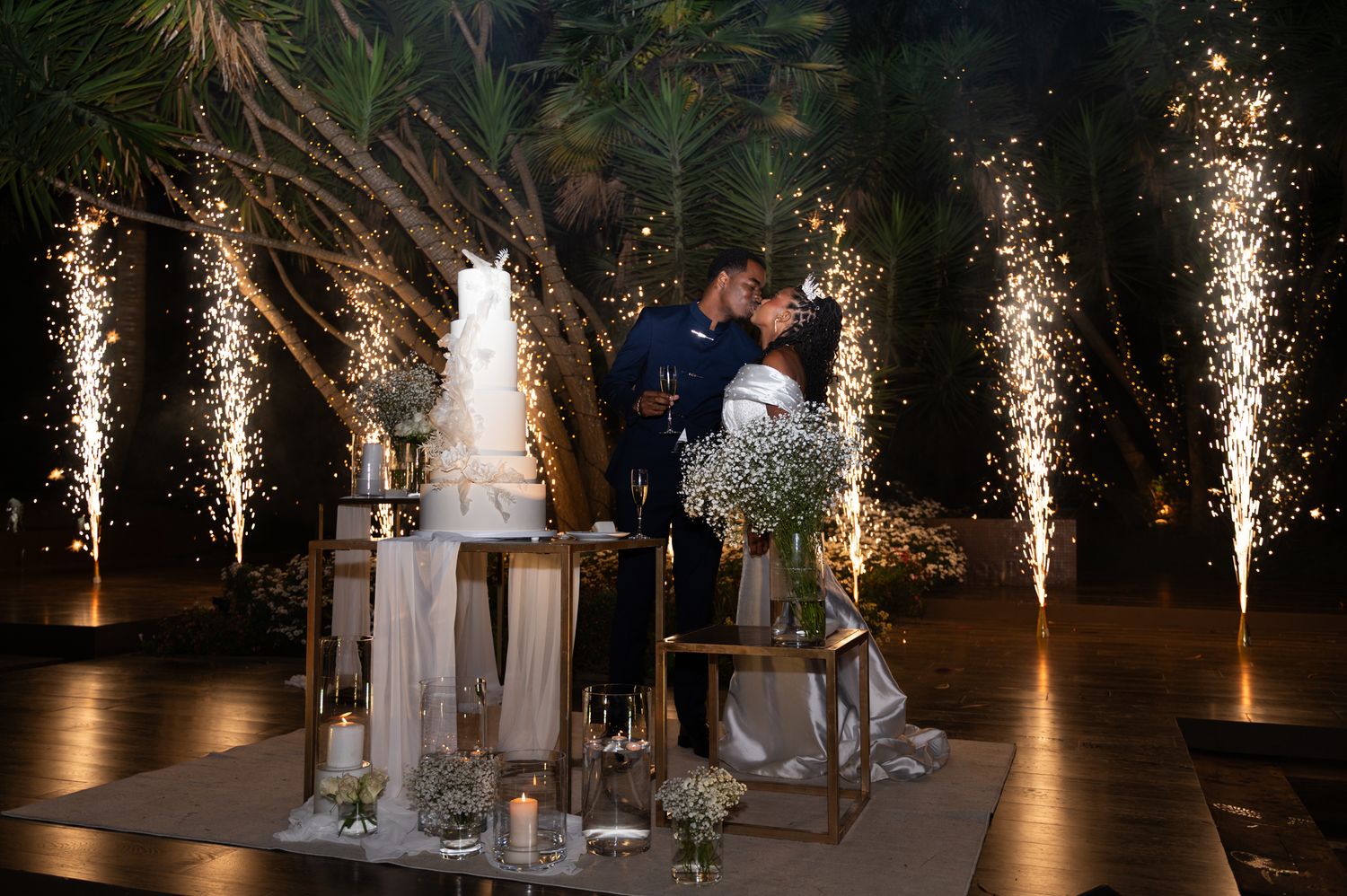 Bride and groom kiss behind wedding cake with dramatic pyrotechnic fountains at outdoor night reception.