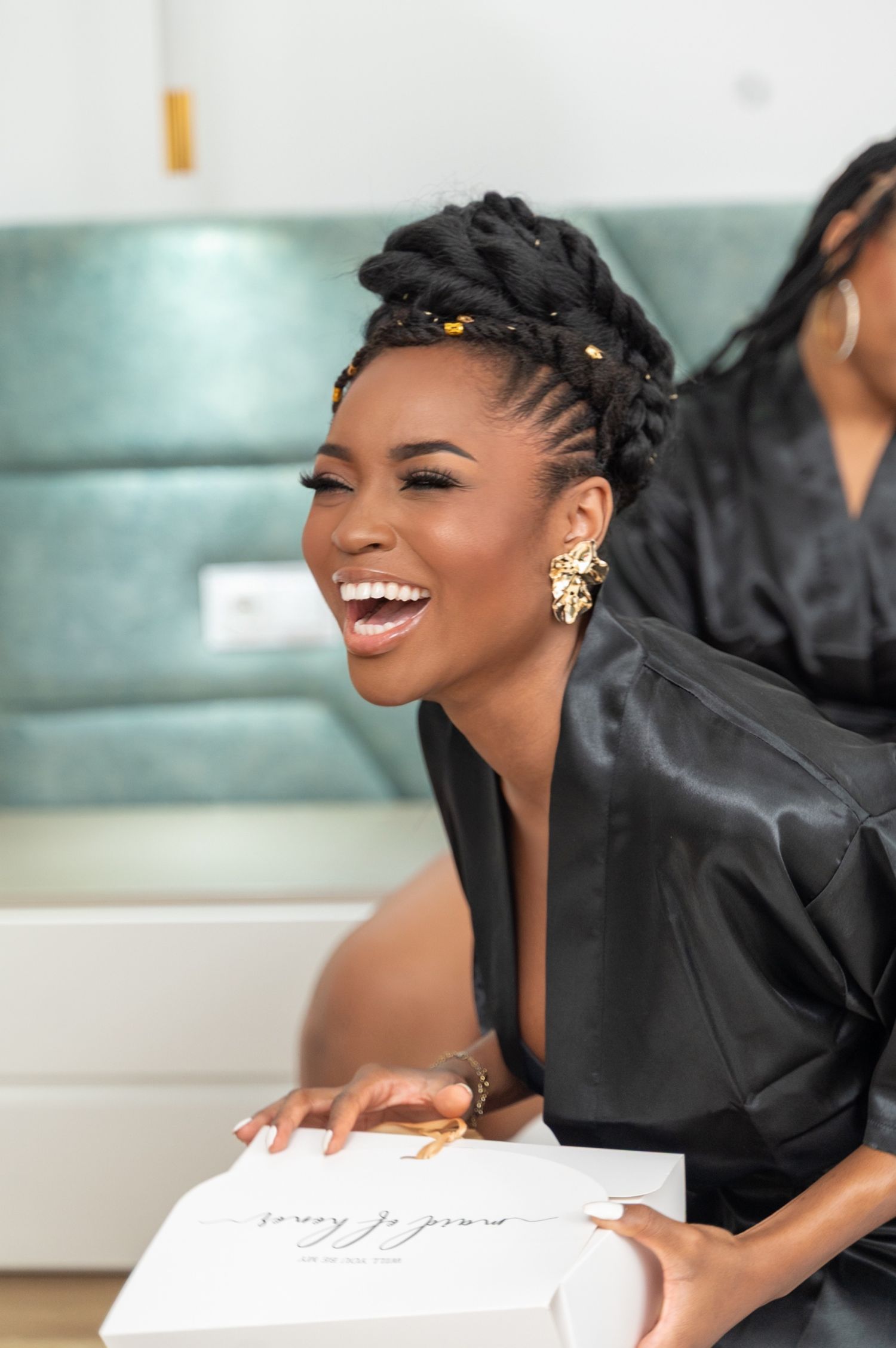 Laughing woman in black satin robe holding a maid of honor gift box with gold earrings.