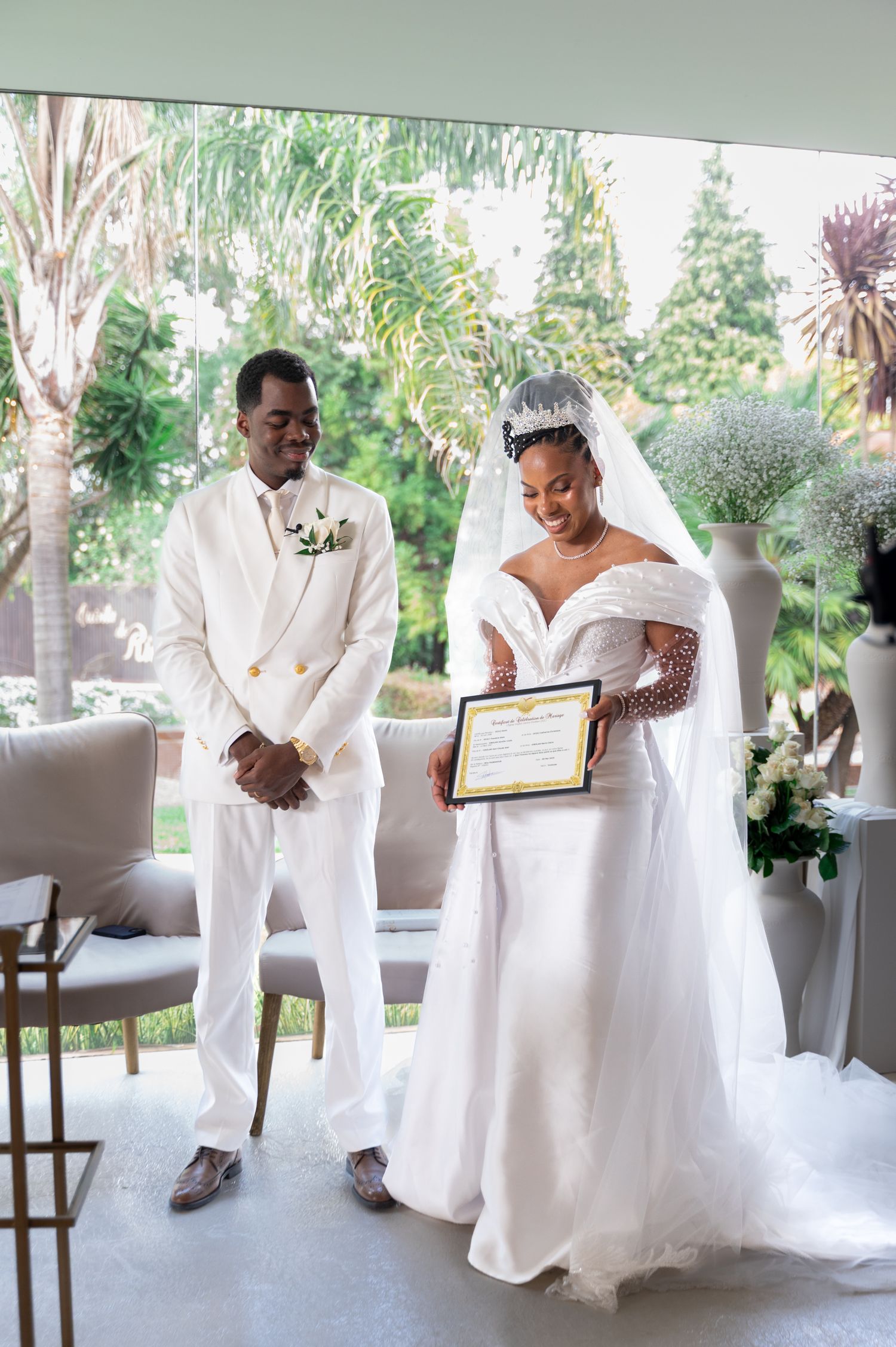 Bride holds marriage certificate beside groom in white suit at elegant wedding ceremony.