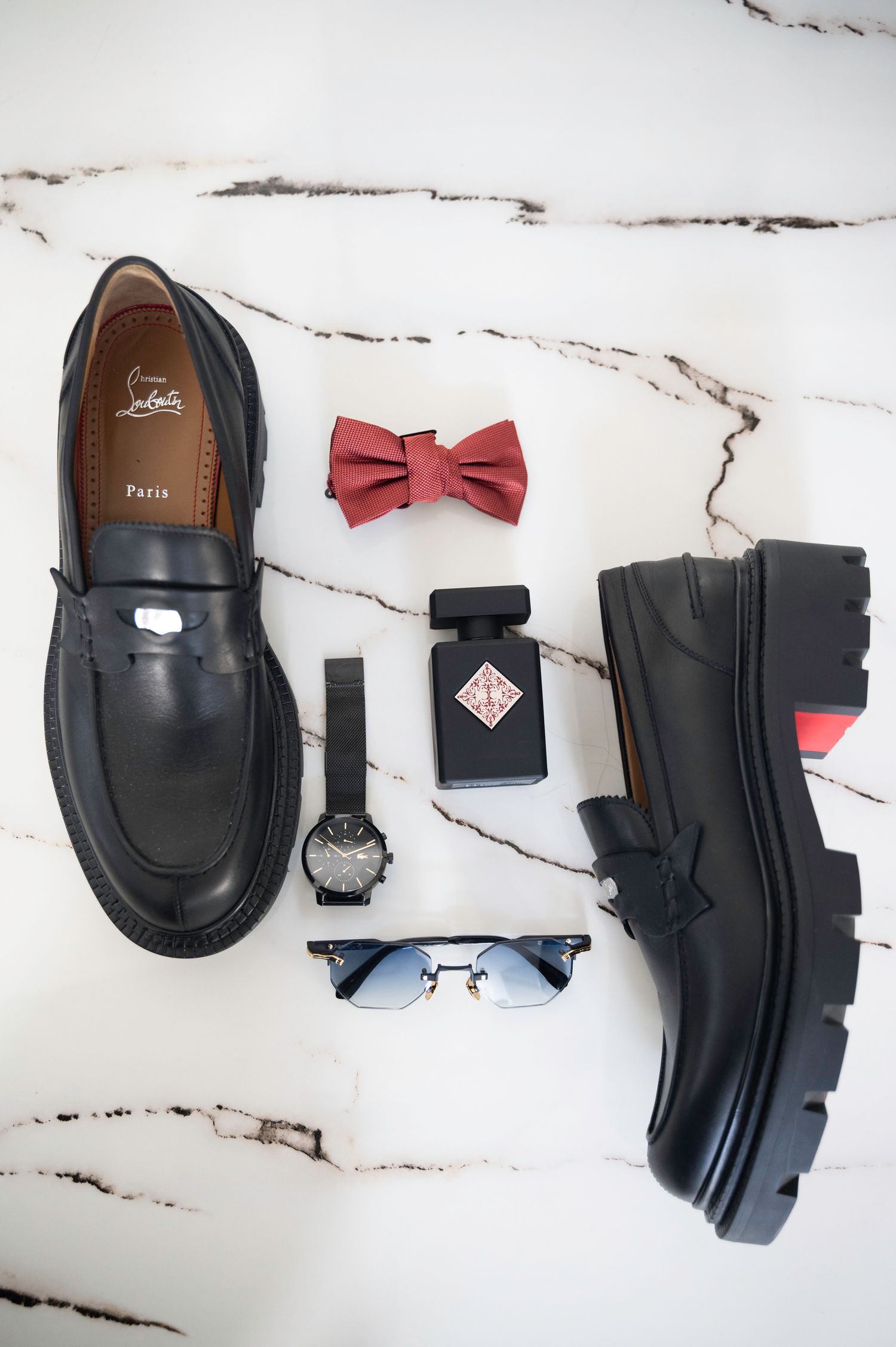 Flat lay of Christian Louboutin loafers, black watch, red bow tie, perfume, and sunglasses on marble.