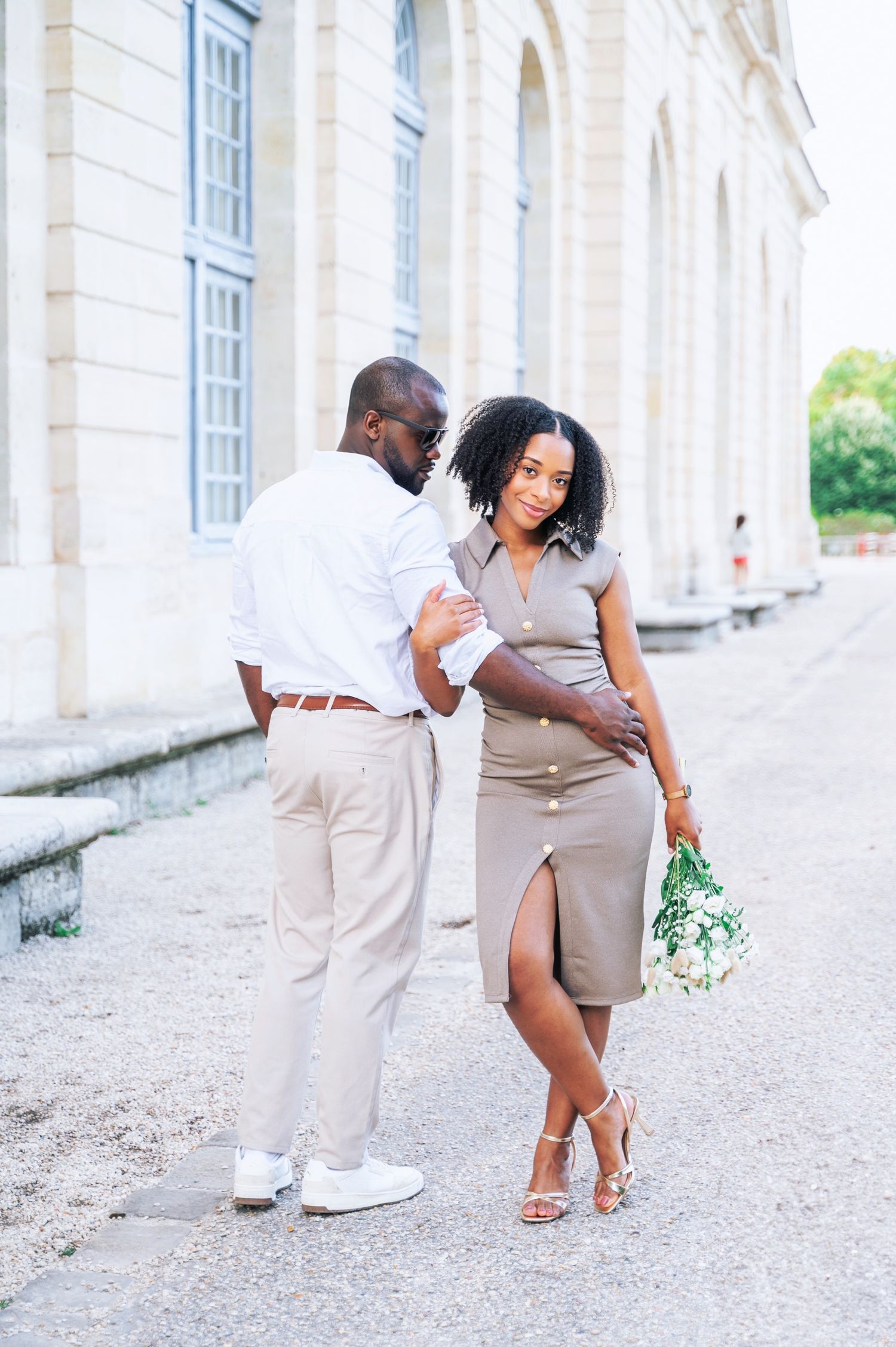 A couple shares a playful moment outside a grand French building, the woman in a taupe dress holding a bouquet.