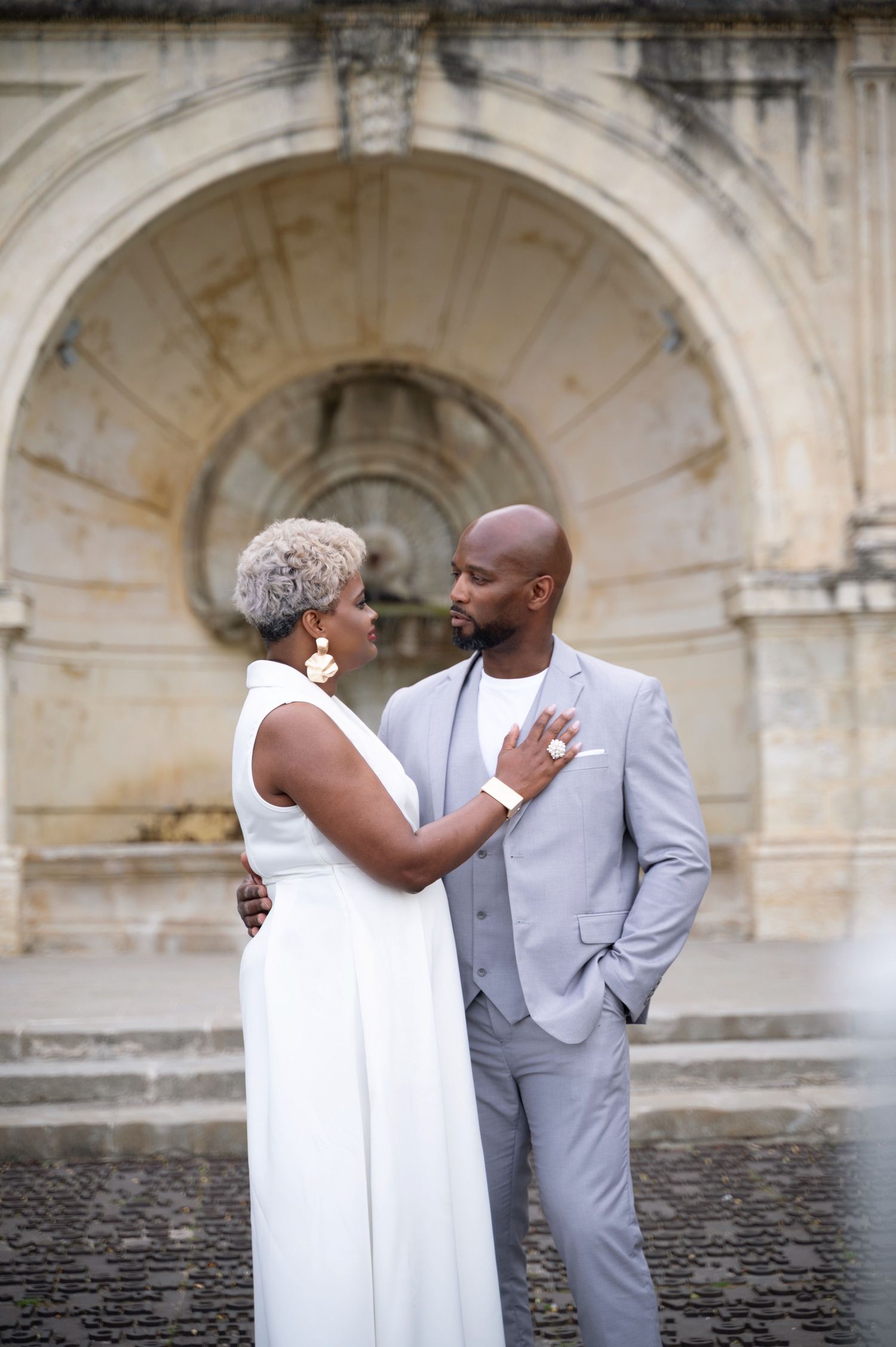 Elegant couple in white and gray attire share an intimate moment by stone fountain.