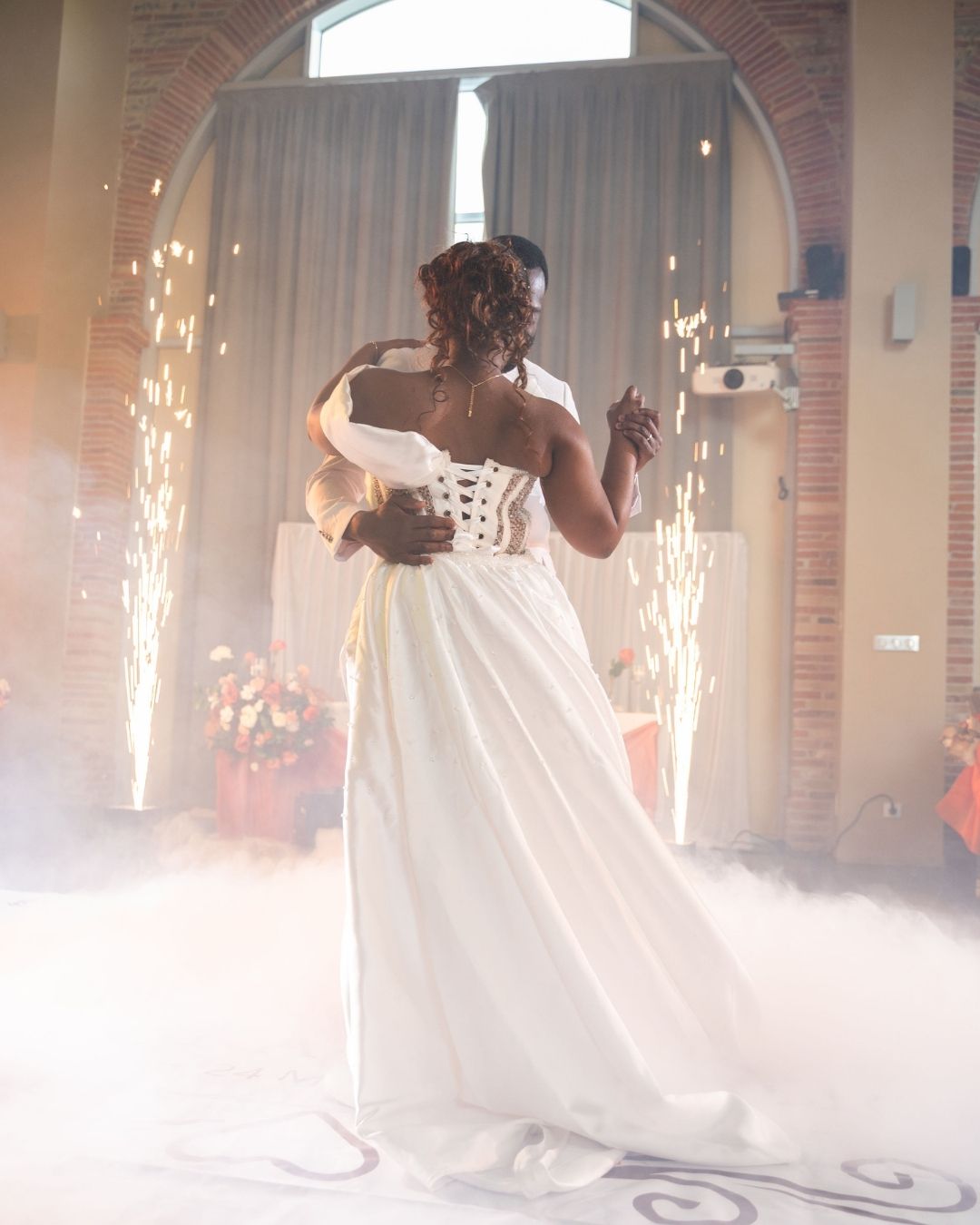 Bride in a corset-back white gown dancing with groom amid fog and sparkling fireworks at wedding reception.