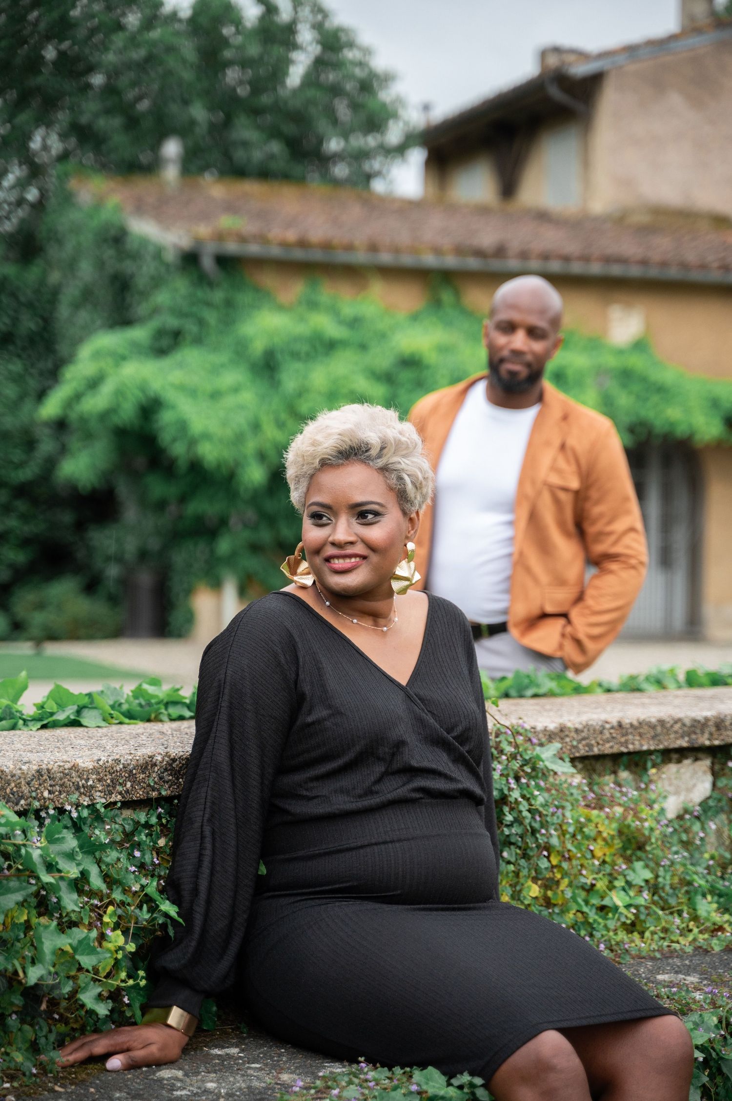 Black woman in black dress with gold earrings sitting on stone wall with man in orange blazer behind her.