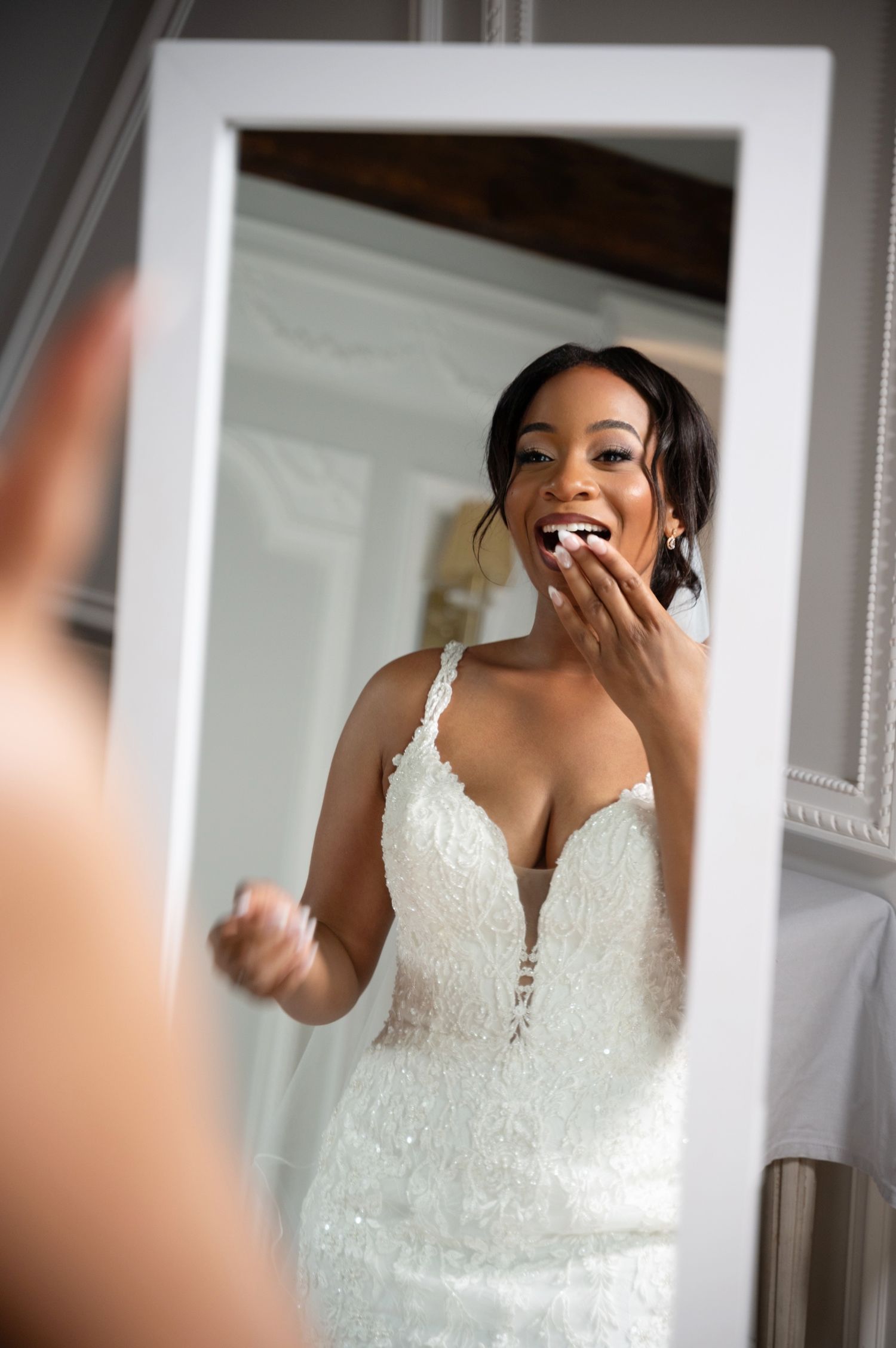A joyful Black bride in a beaded white gown laughs at her reflection in a white framed mirror.