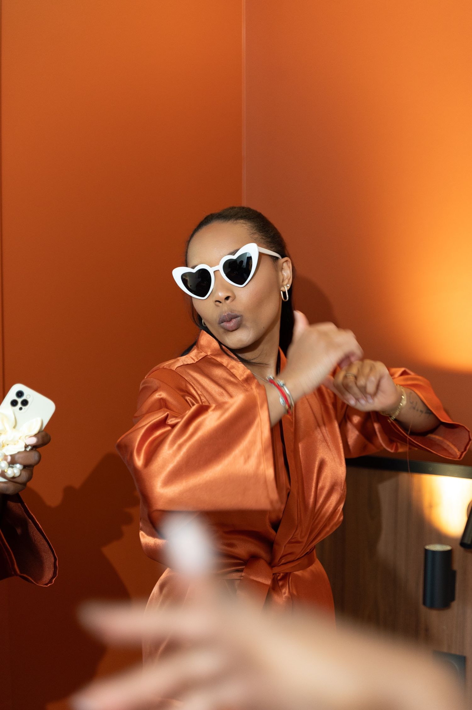 A bridesmaid in an orange satin robe wears white heart-shaped sunglasses and dances playfully before the wedding.