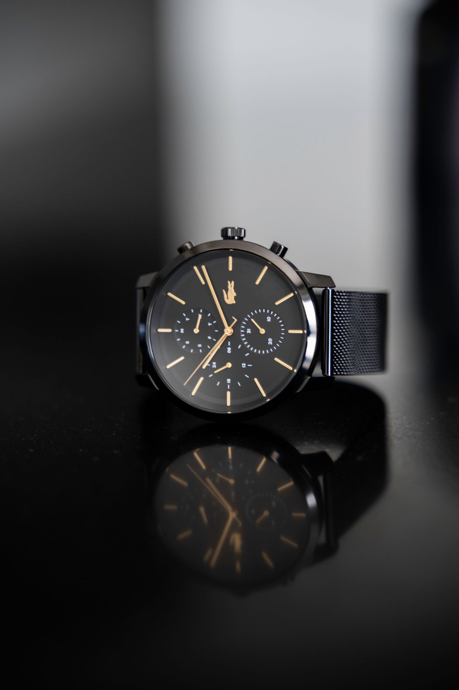 Lacoste black chronograph watch with gold accents and mesh bracelet on reflective dark surface.