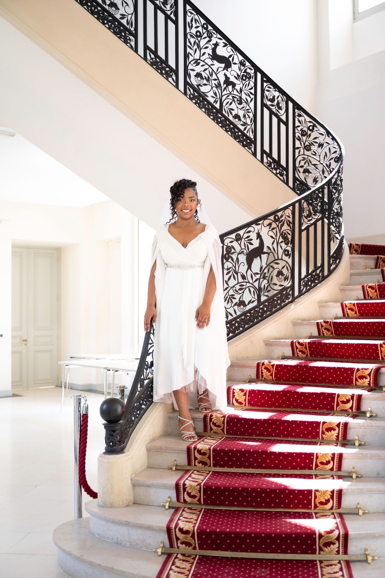 Elegant Black bride in white gown descends grand staircase with red carpet runner.