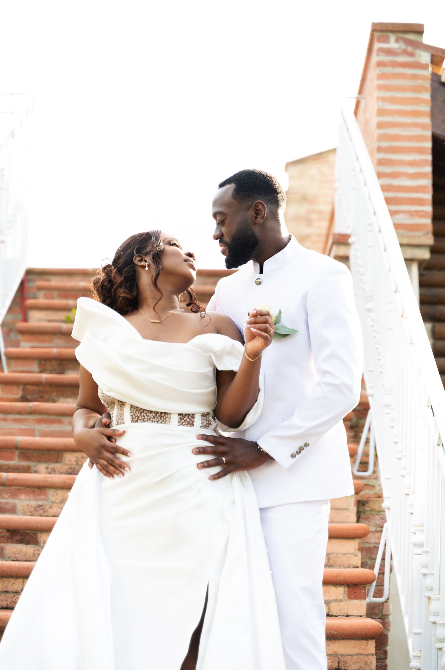 Bride and groom in white wedding outfits sharing a romantic gaze on sunlit brick staircase.