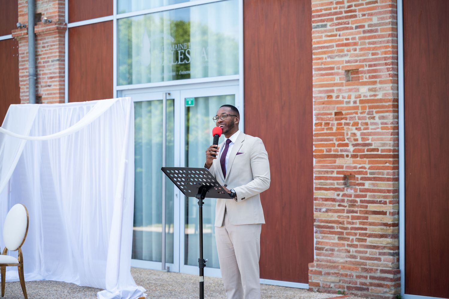 Officiant in beige suit speaks at podium during outdoor wedding ceremony near brick building entrance.
