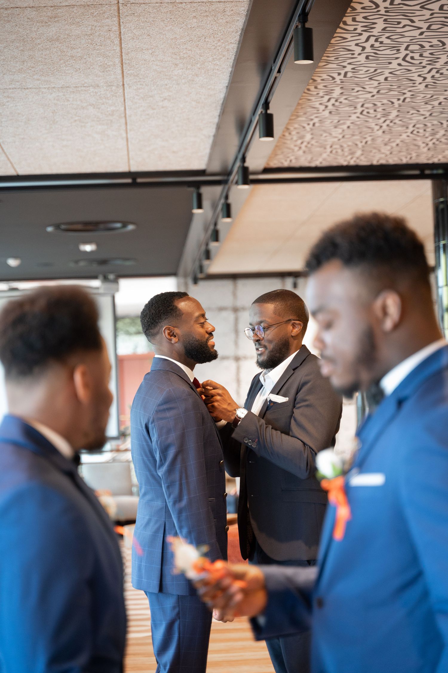 Groomsman helping groom with bow tie while other groomsmen in blue suits stand nearby in modern venue.