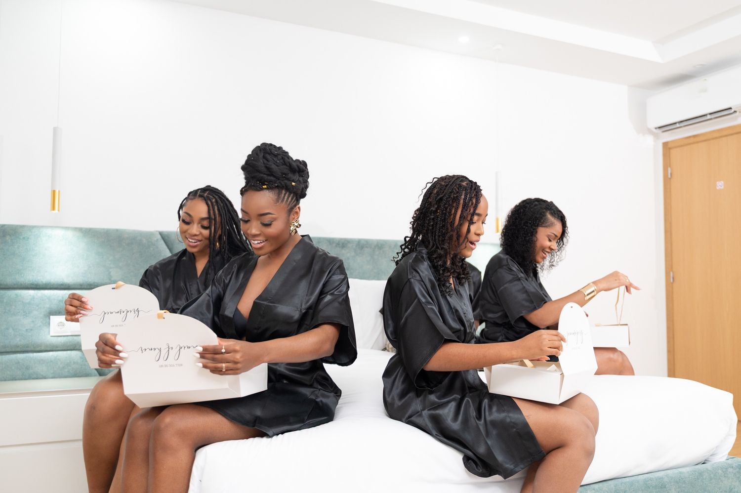 Four bridesmaids in black satin robes sitting on a bed opening gift boxes together.