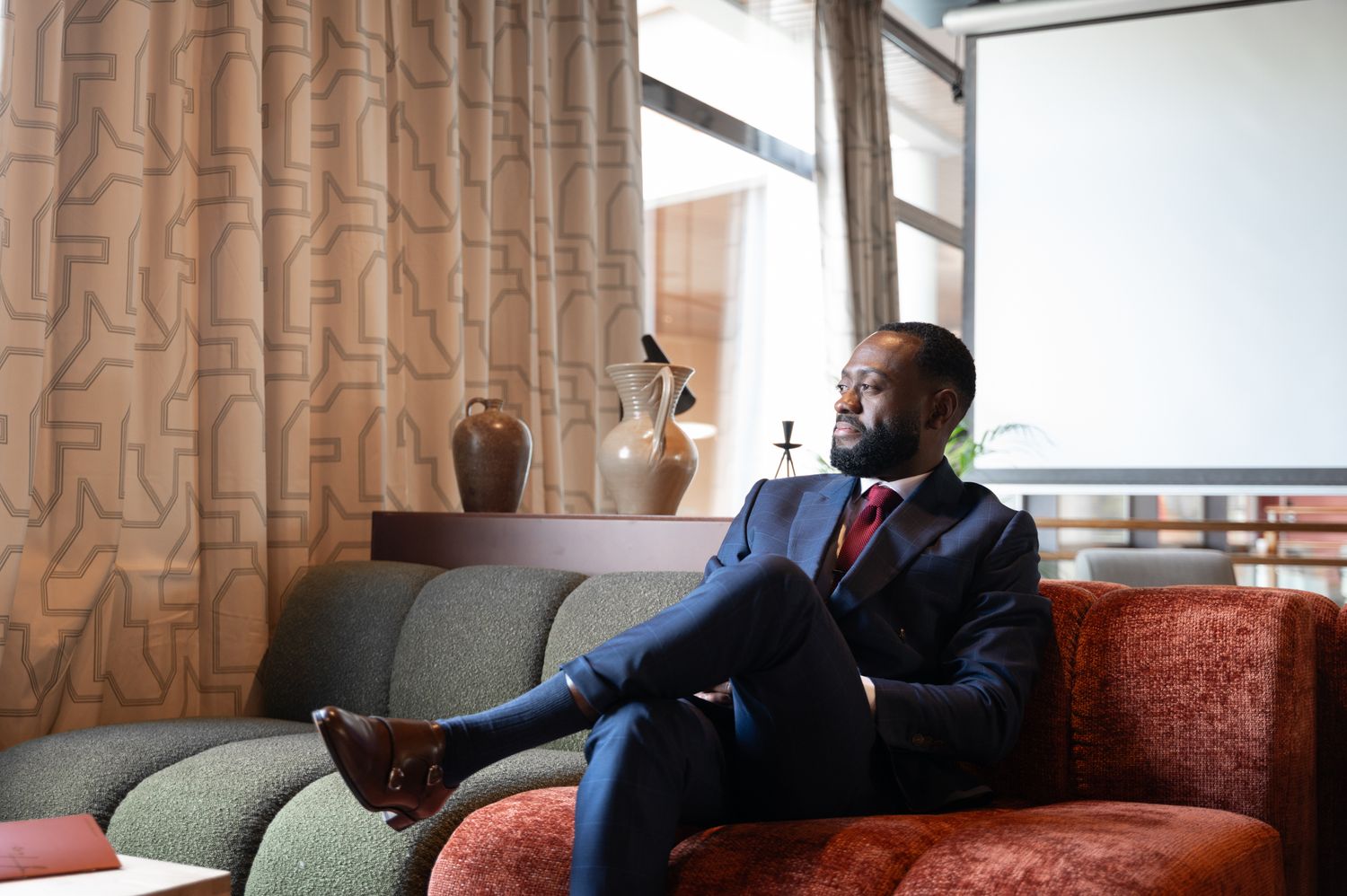 A smiling Black man in a navy suit sits on a couch in an elegantly decorated room.