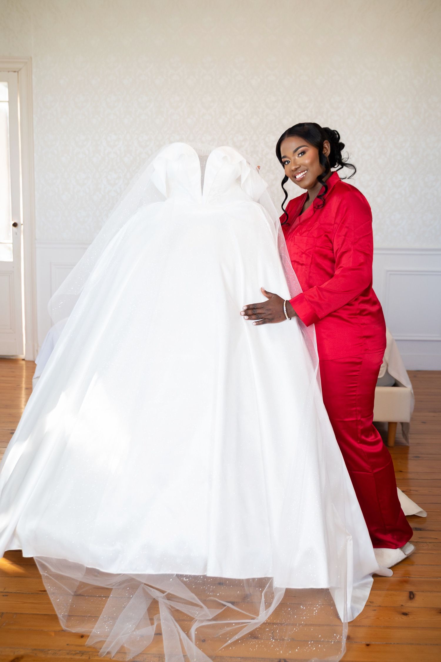 A smiling bride in red satin pajamas lovingly embraces her elegant white ball gown wedding dress in a bright room.