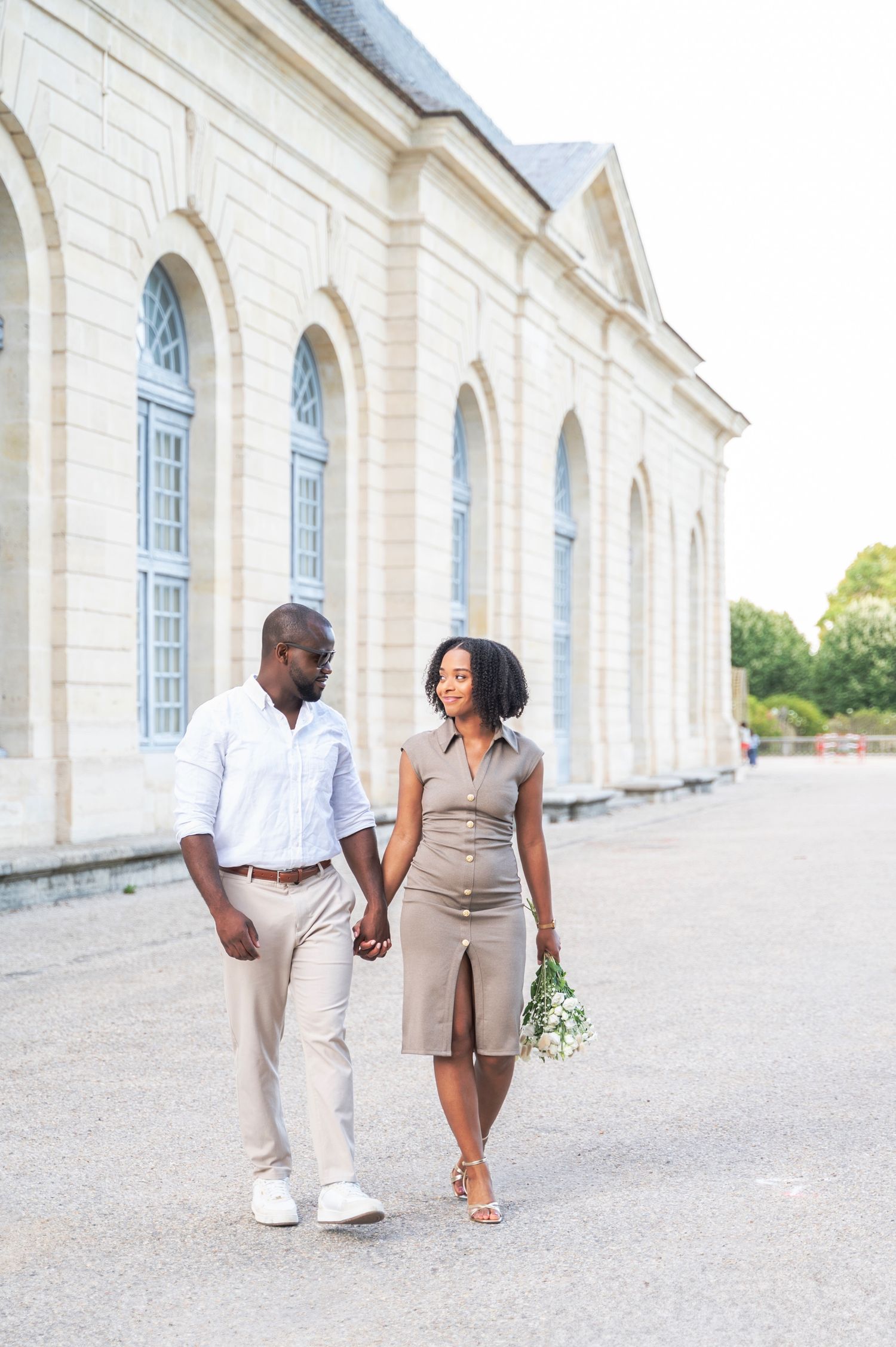 A stylish couple strolls hand in hand along a French palace exterior, the woman smiling and holding flowers.