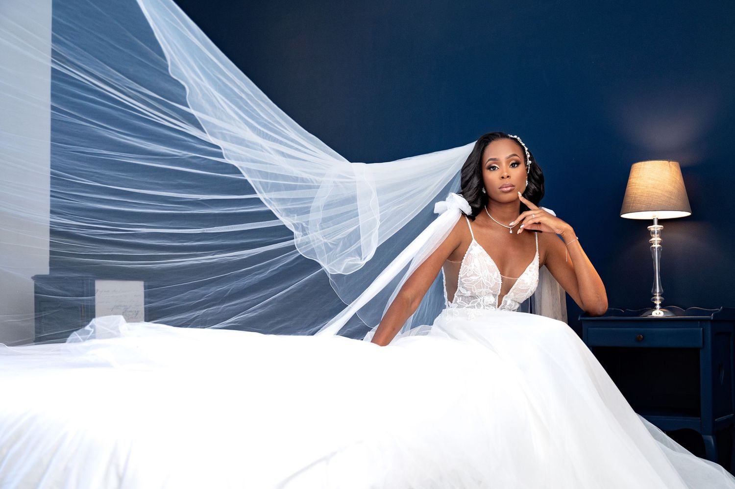 Bride in a white wedding gown with flowing veil sits on a bed against a deep blue wall.