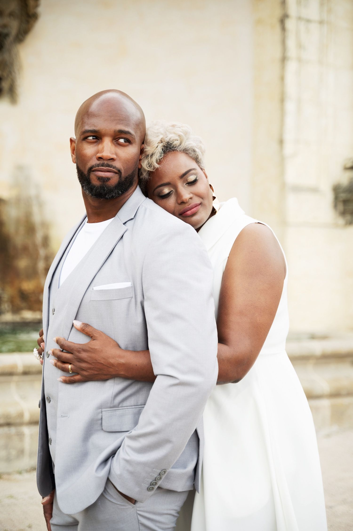 Black couple in elegant embrace, woman in white dress leaning on man in light gray suit outdoors.