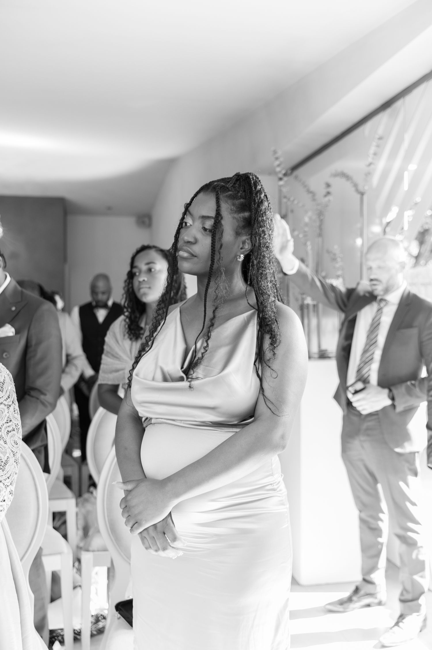 Emotional pregnant wedding guest in white dress stands with eyes closed during ceremony.