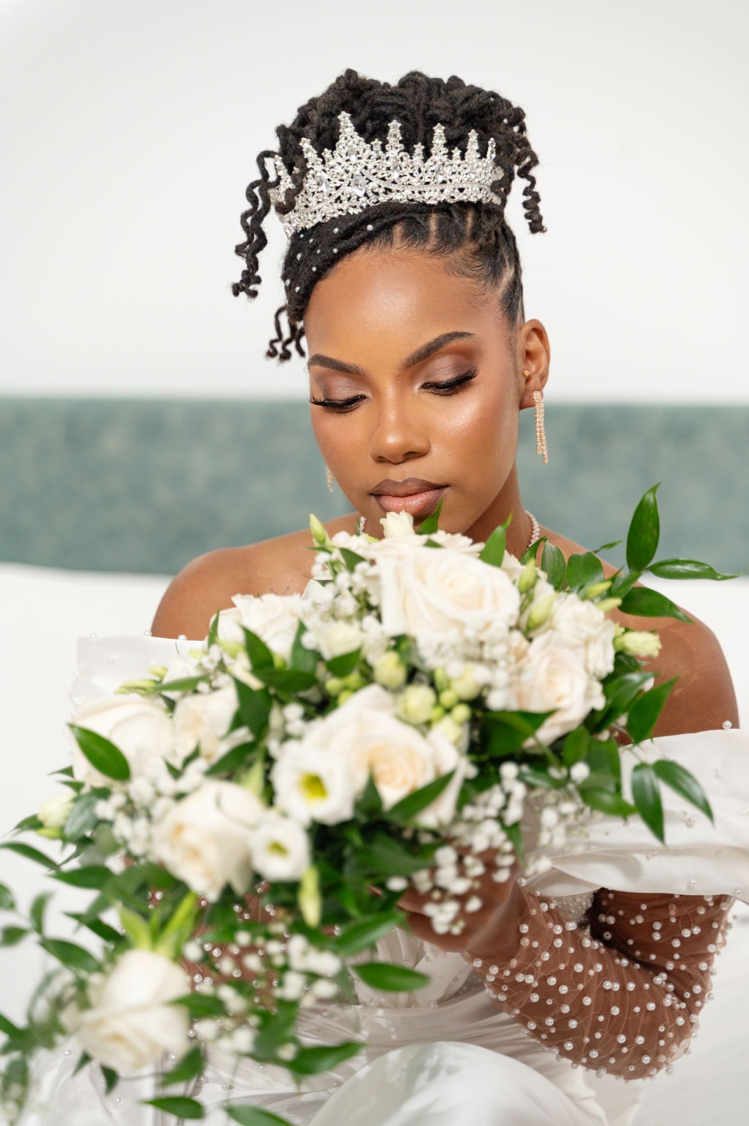 Black bride in white gown and tiara gazes at bouquet of white roses and greenery.