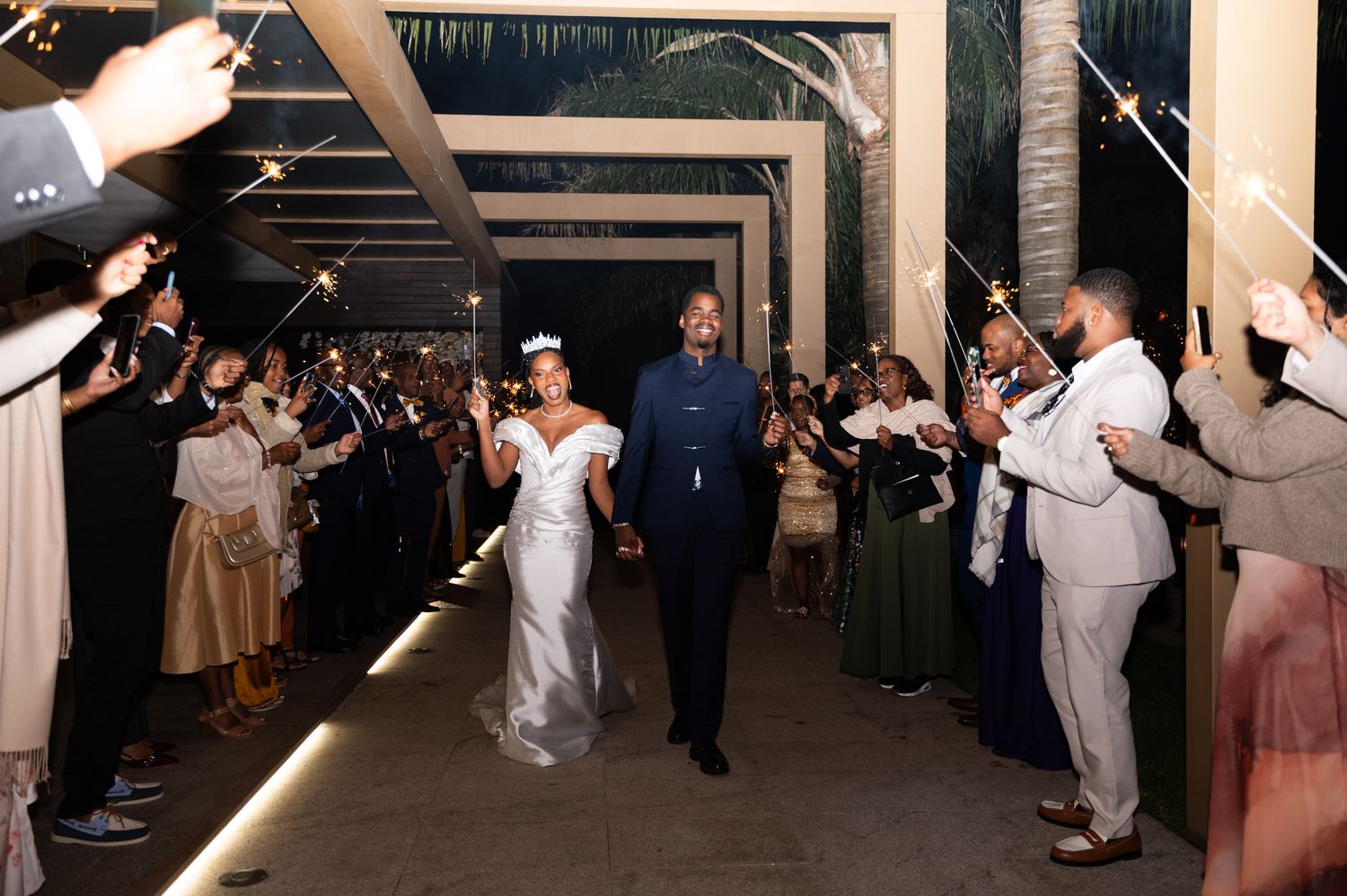 Newlyweds walk through sparkler exit tunnel at night surrounded by cheering wedding guests outdoors.