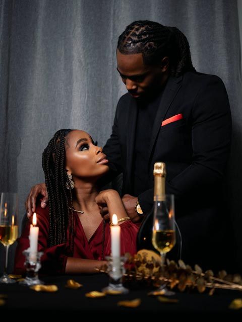 Elegant couple at romantic candlelit dinner, woman in red dress seated as man in black suit stands close.
