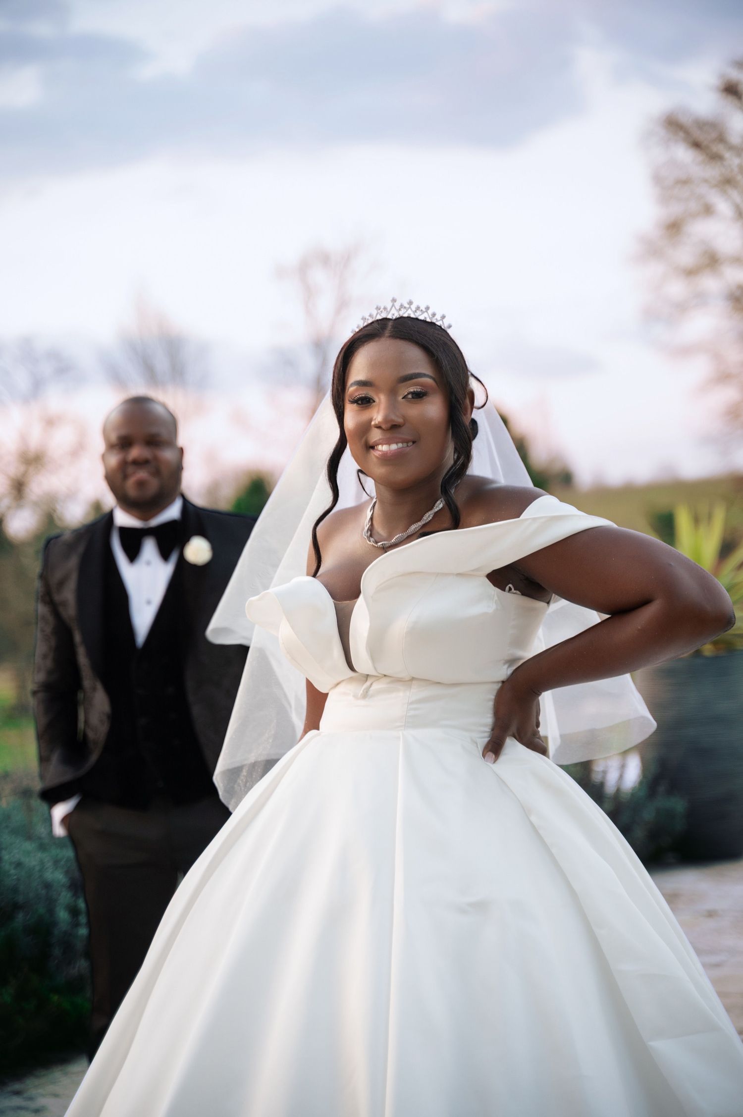 Elegant Black bride in off-shoulder ball gown with tiara and veil outdoors.