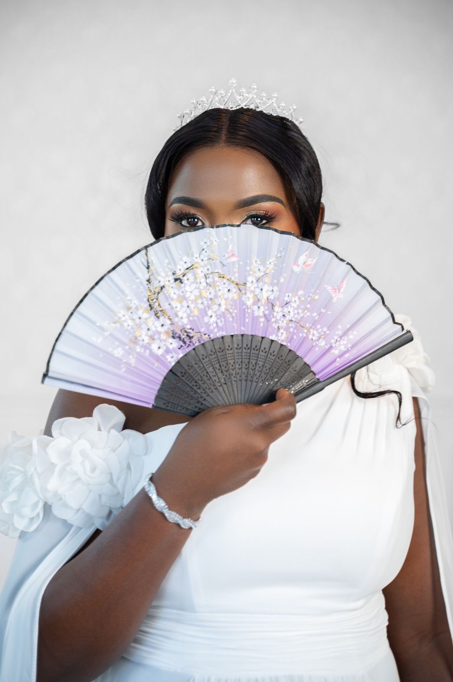 A Black bride in a white gown and crystal tiara playfully hides her face behind a purple floral hand fan.