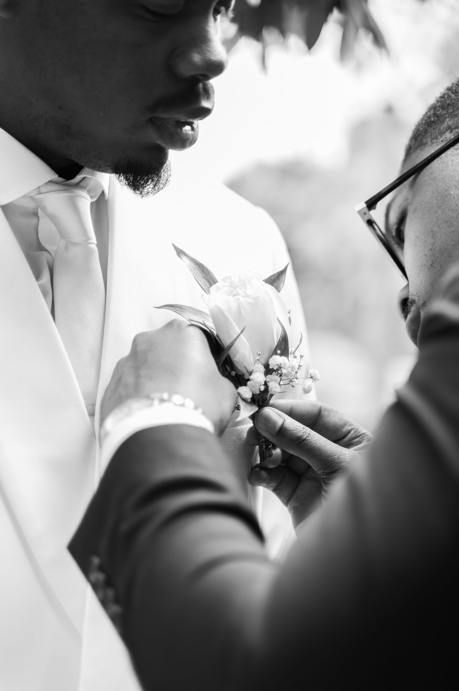 Black and white close-up of groomsman pinning floral boutonniere on groom's white suit.