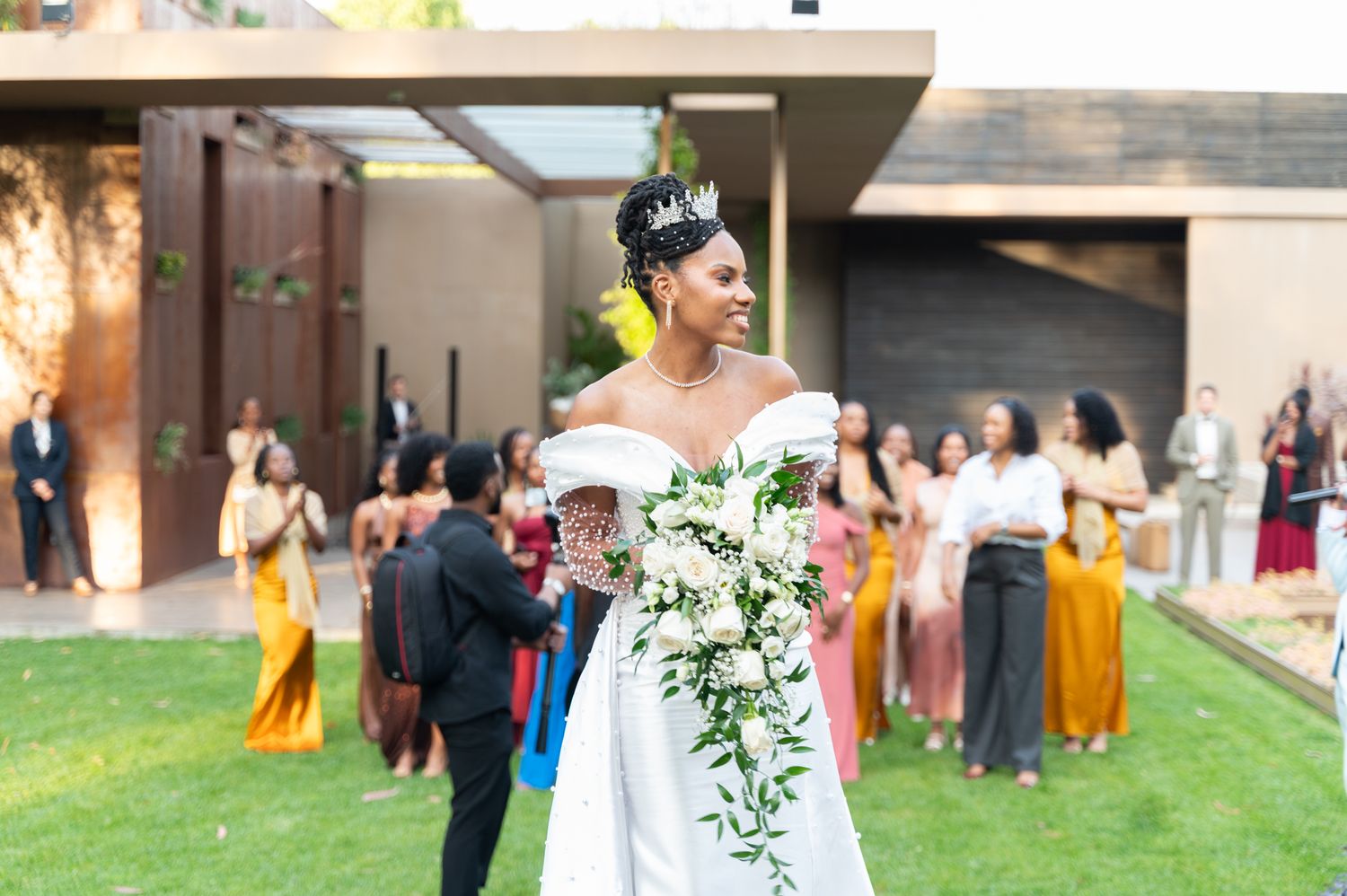 Elegant bride wearing crown and off-shoulder white gown holds cascading floral bouquet at outdoor wedding.